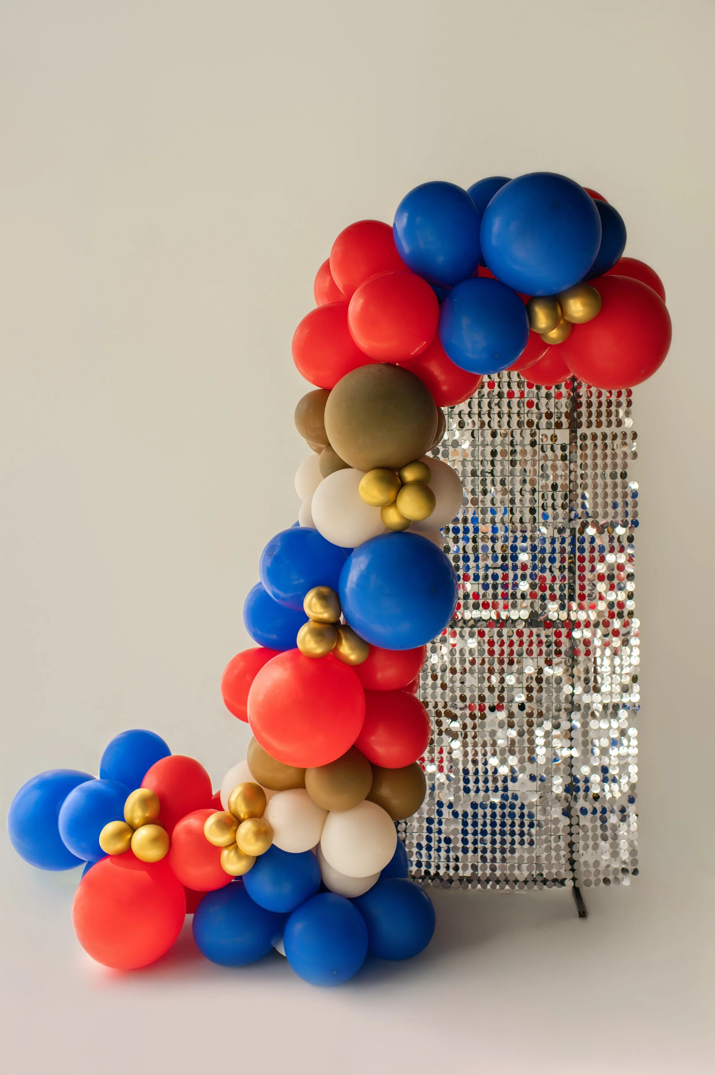 Arrangement of colorful balloons in red, blue, white, gold, and beige, with a backdrop of sequin panels.