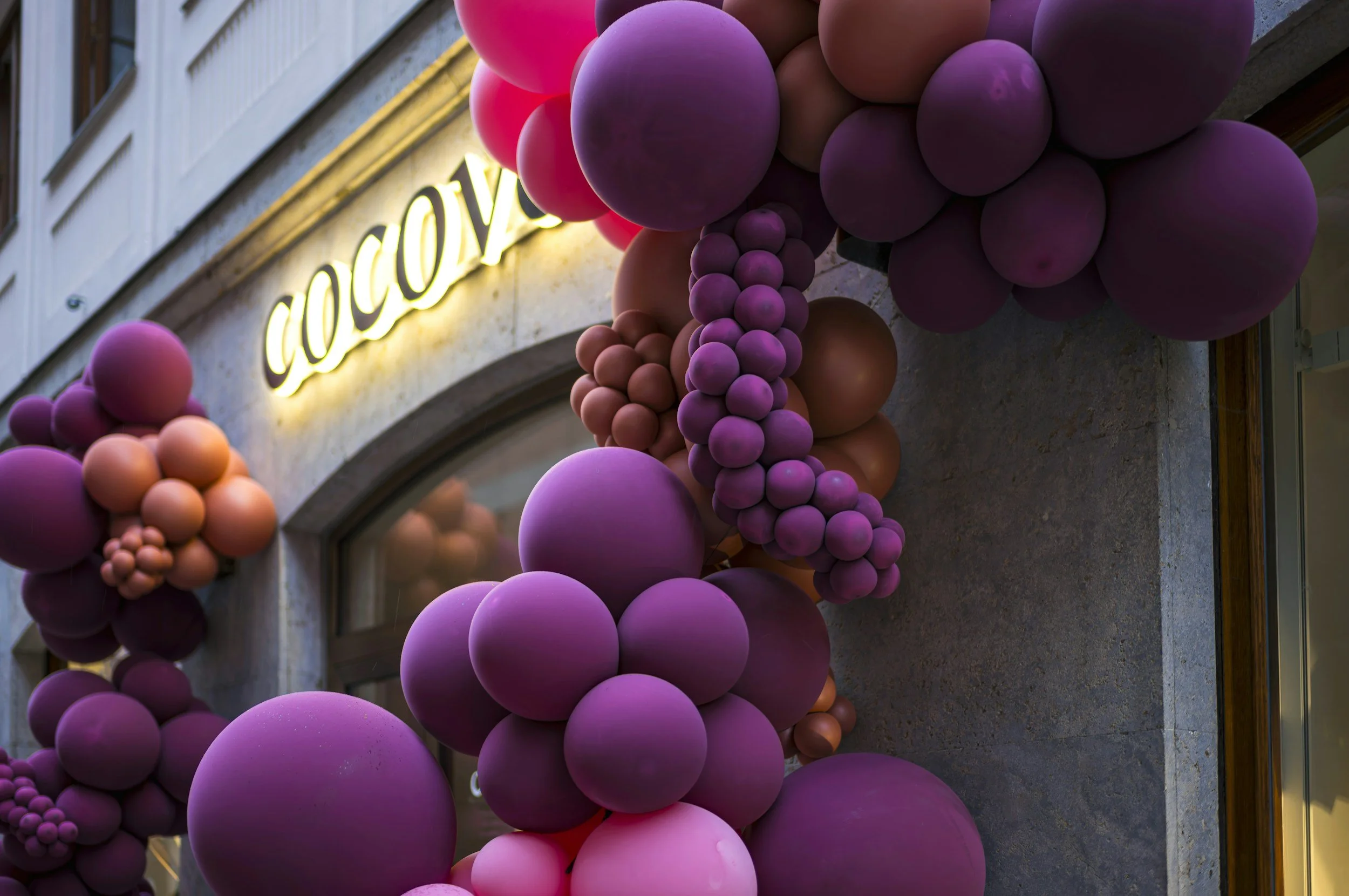Decorative purple and pink balloons arranged along a building facade with a neon sign in the background.