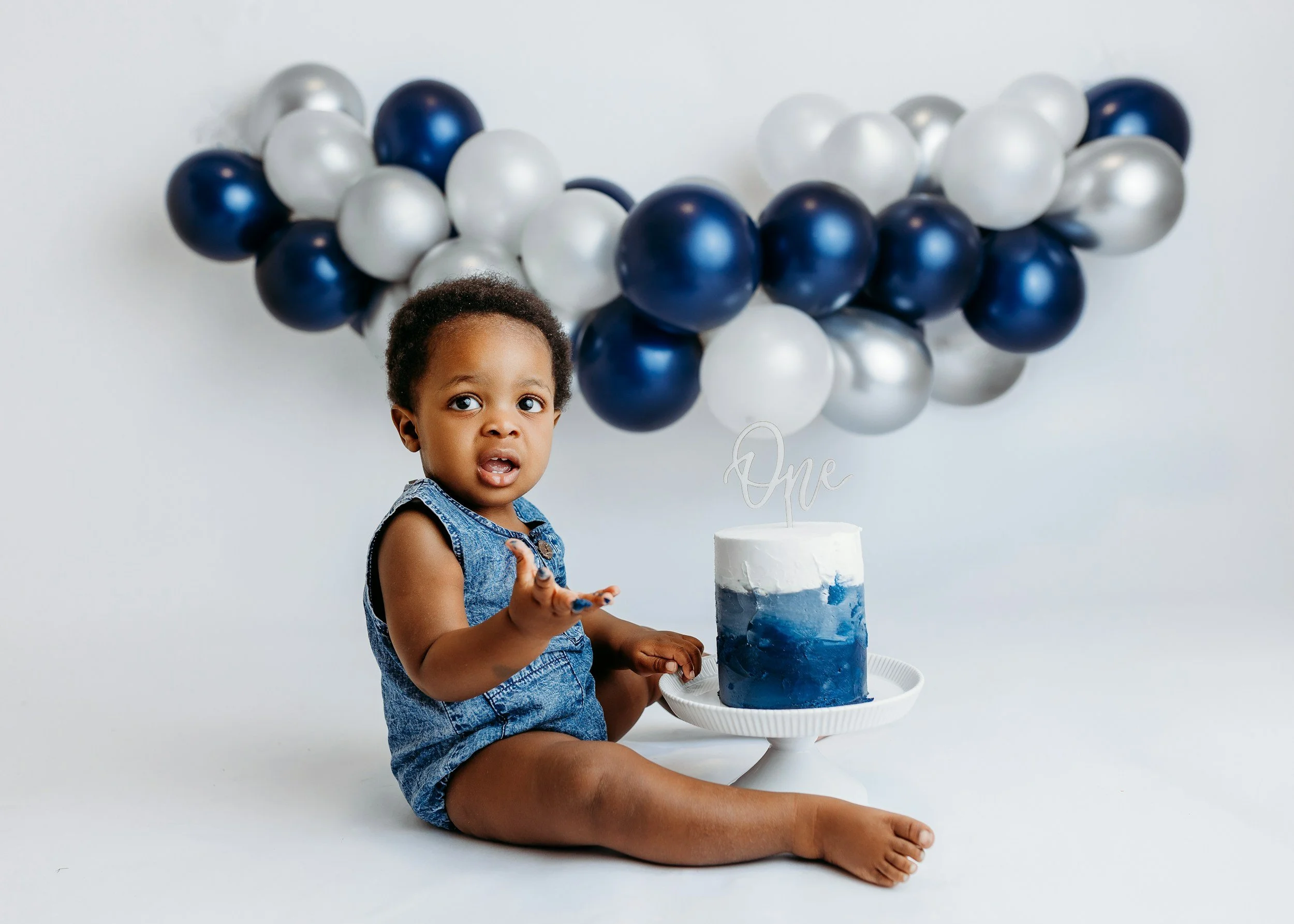 A young child with curly hair and brown skin sitting on the floor in front of a white cake decorated with blue and white frosting. The child is wearing a denim outfit and holding their hand out. Behind the cake is a large cluster of blue, white, and silver balloons arranged in a semi-circle, with a white wall as the background.