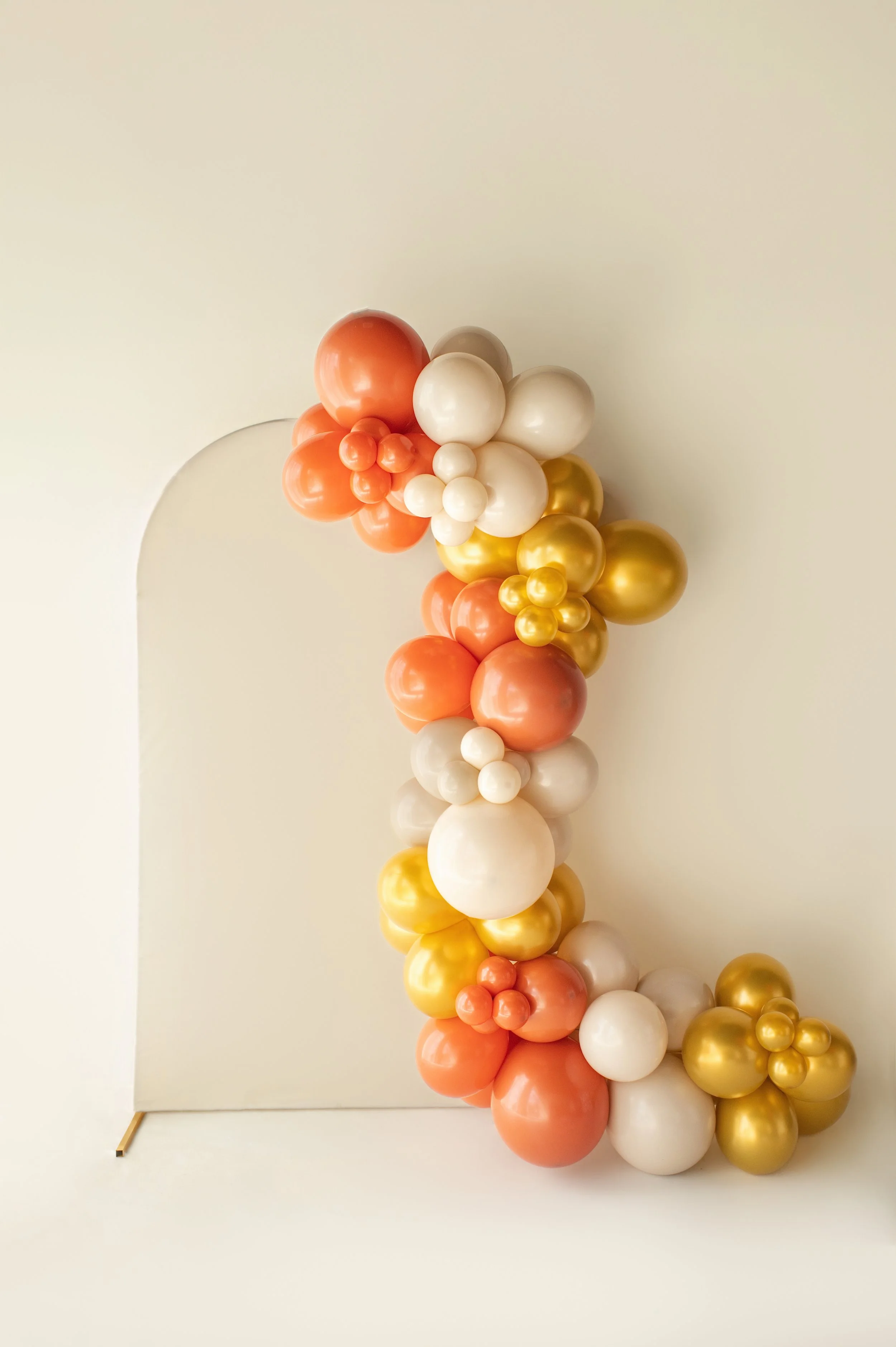 Colorful balloon garland in shades of white, gold, peach, and coral arranged along a white wall with an arch-shaped mirror.