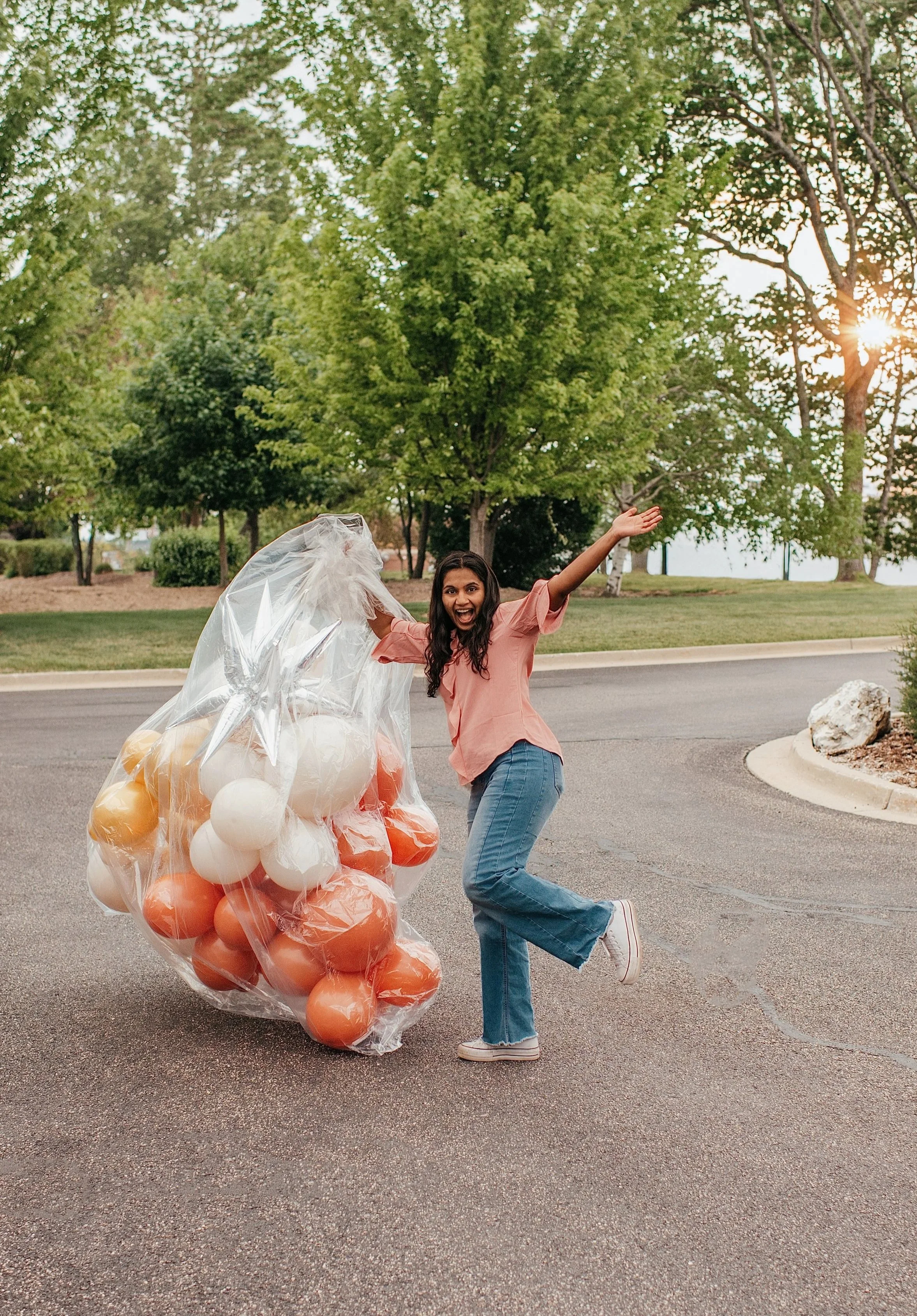 A woman smiling and jumping joyfully while holding balloons, standing outdoors on a paved road with green trees and the setting sun in the background.