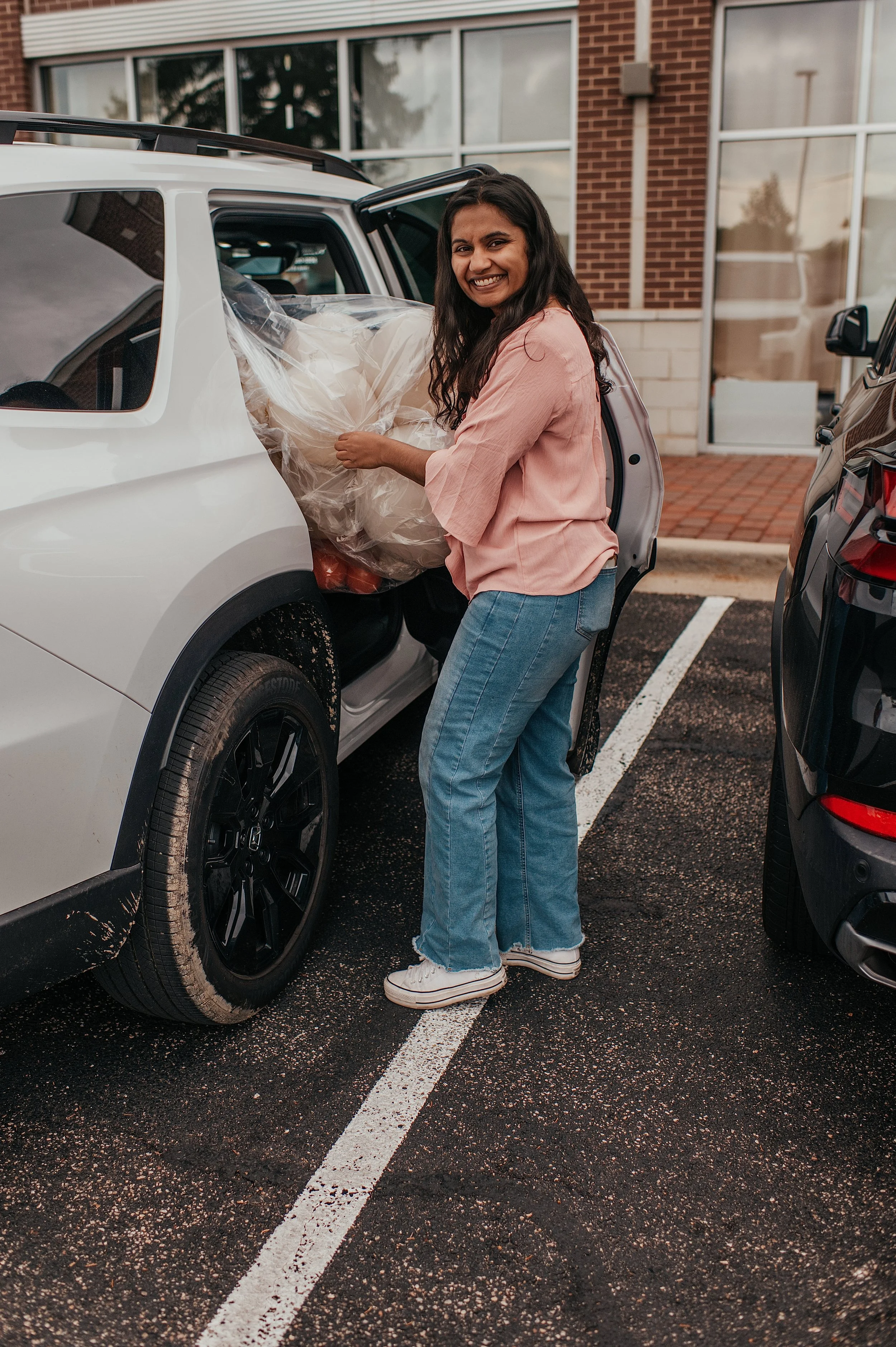 A woman with long dark hair, wearing a pink shirt, blue jeans, and white sneakers, loading a large wrapped gift into the back seat of a white SUV parked in a parking lot in front of a brick building.