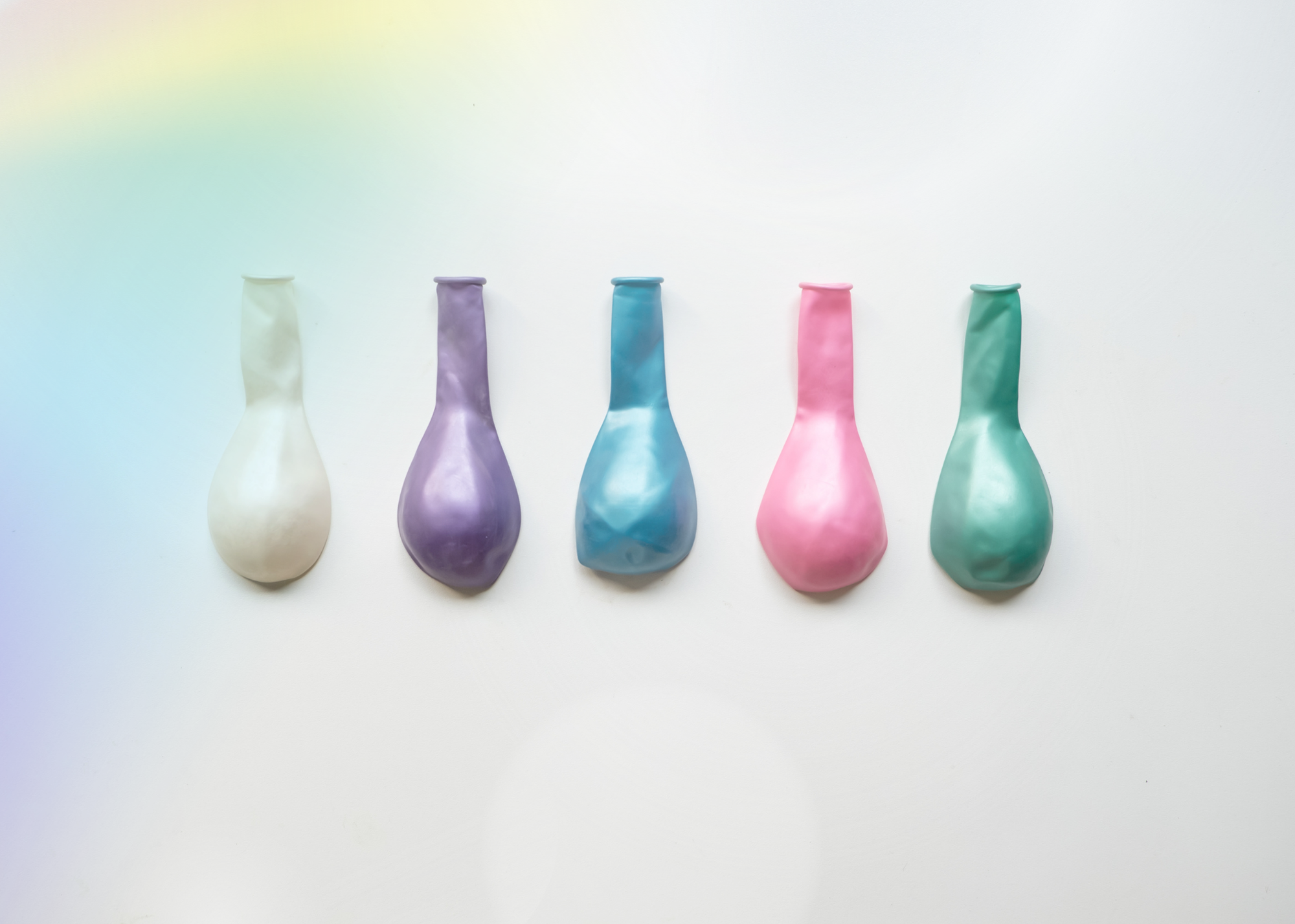 Five deflated pastel-colored balloons in a row on a white surface, with colors from left to right: cream, lavender, blue, pink, and green.