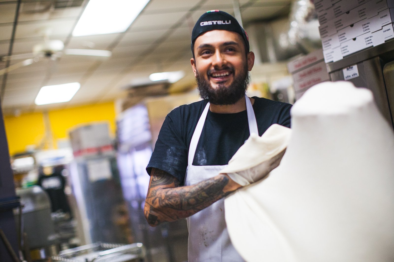 A smiling man wearing a black cap with 'Castelli' written on it, a black t-shirt, and a white apron, working in a kitchen or bakery environment