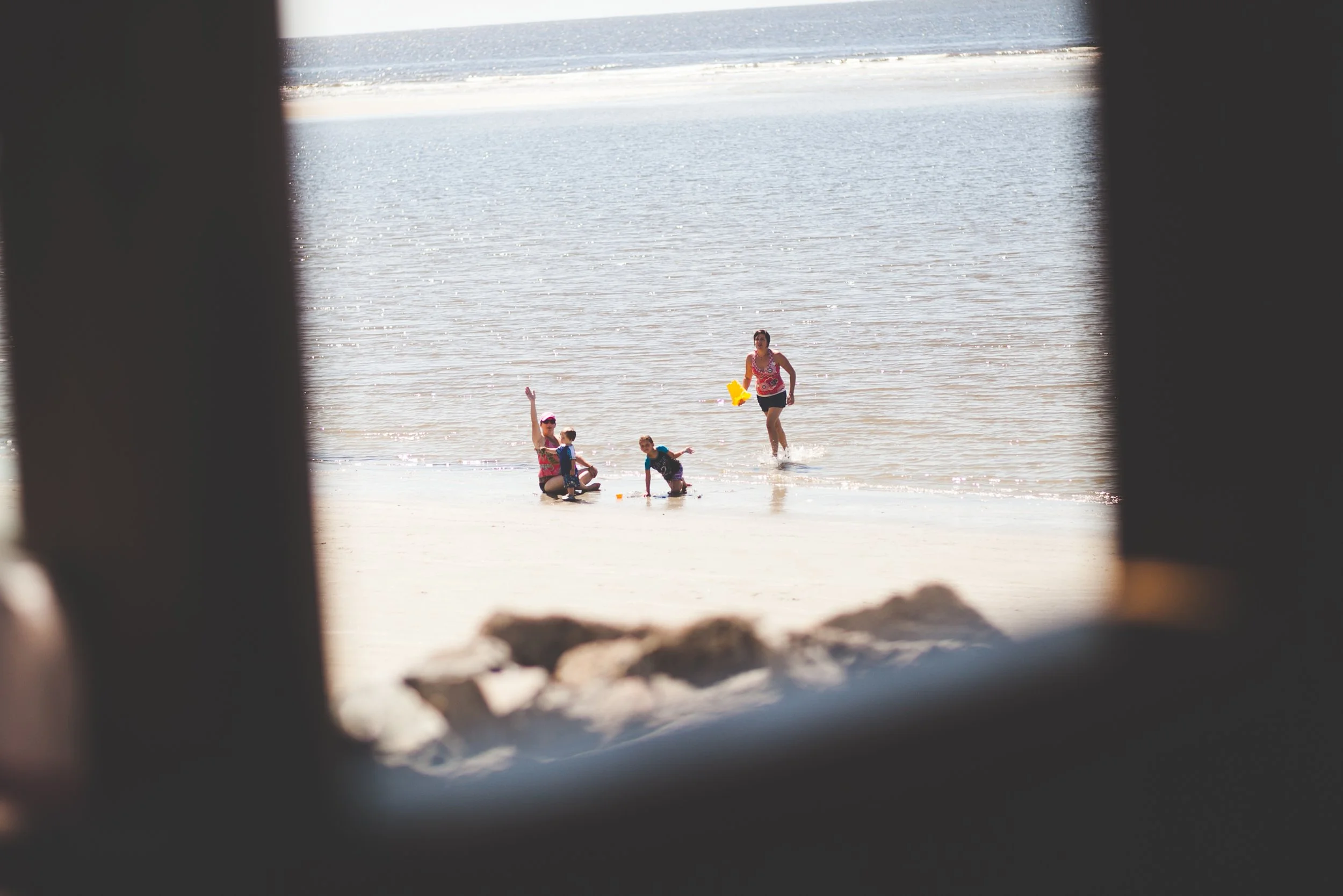 A group of people, including children and a woman, playing and splashing in the shallow water at the beach, viewed through a window or opening with blurred edges.