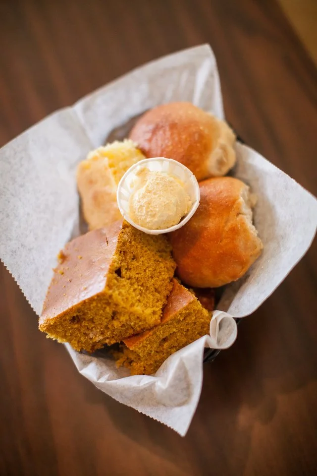 An assortment of banana bread and muffins with a small cup of butter on top, served in a paper-lined basket on a wooden surface.