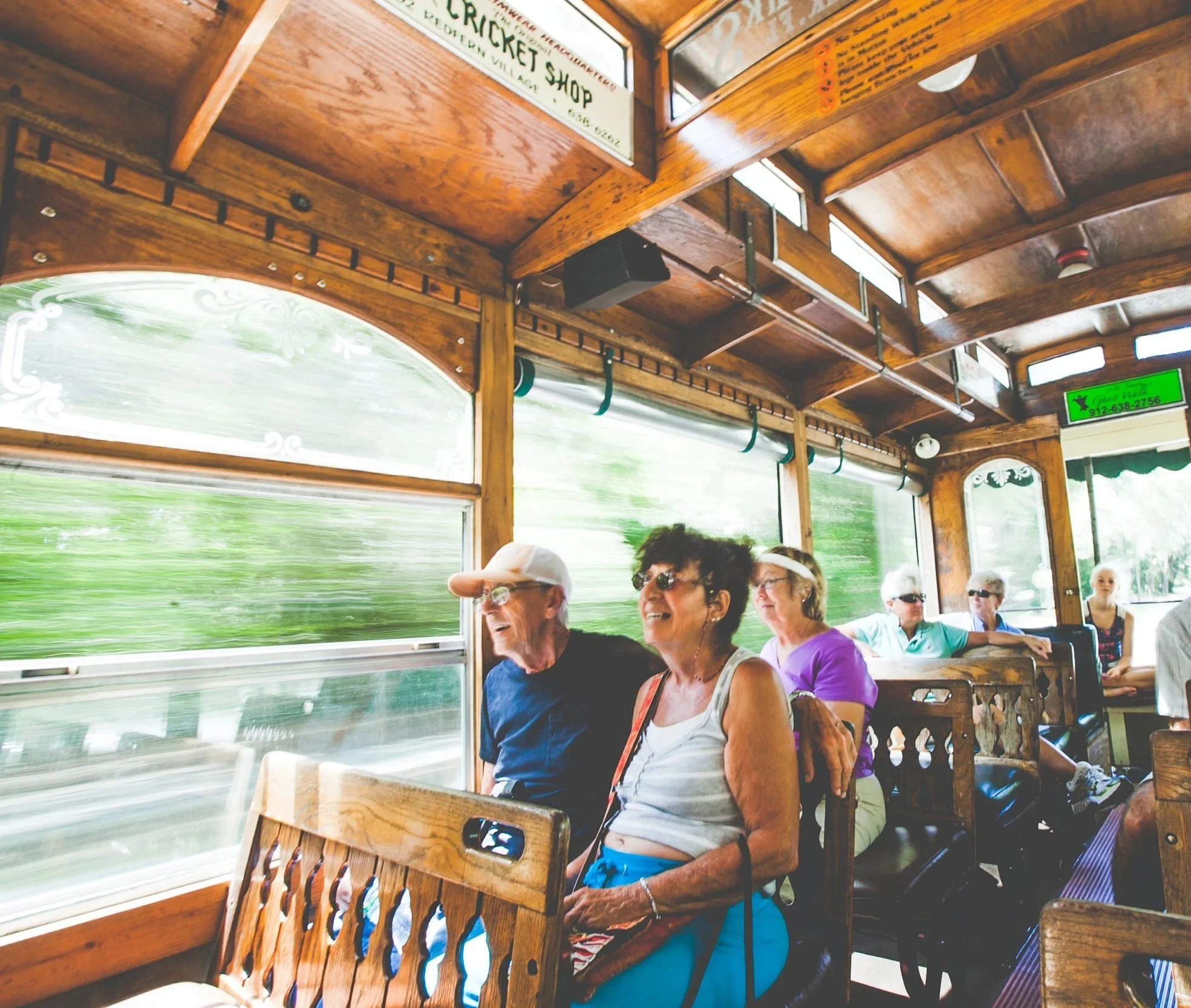 Inside a trolley with wooden interior and windows with green scenery outside. Several older women are sitting, one smiling with sunglasses, and other partially visible women also appear to be seated, enjoying a tour or outing.
