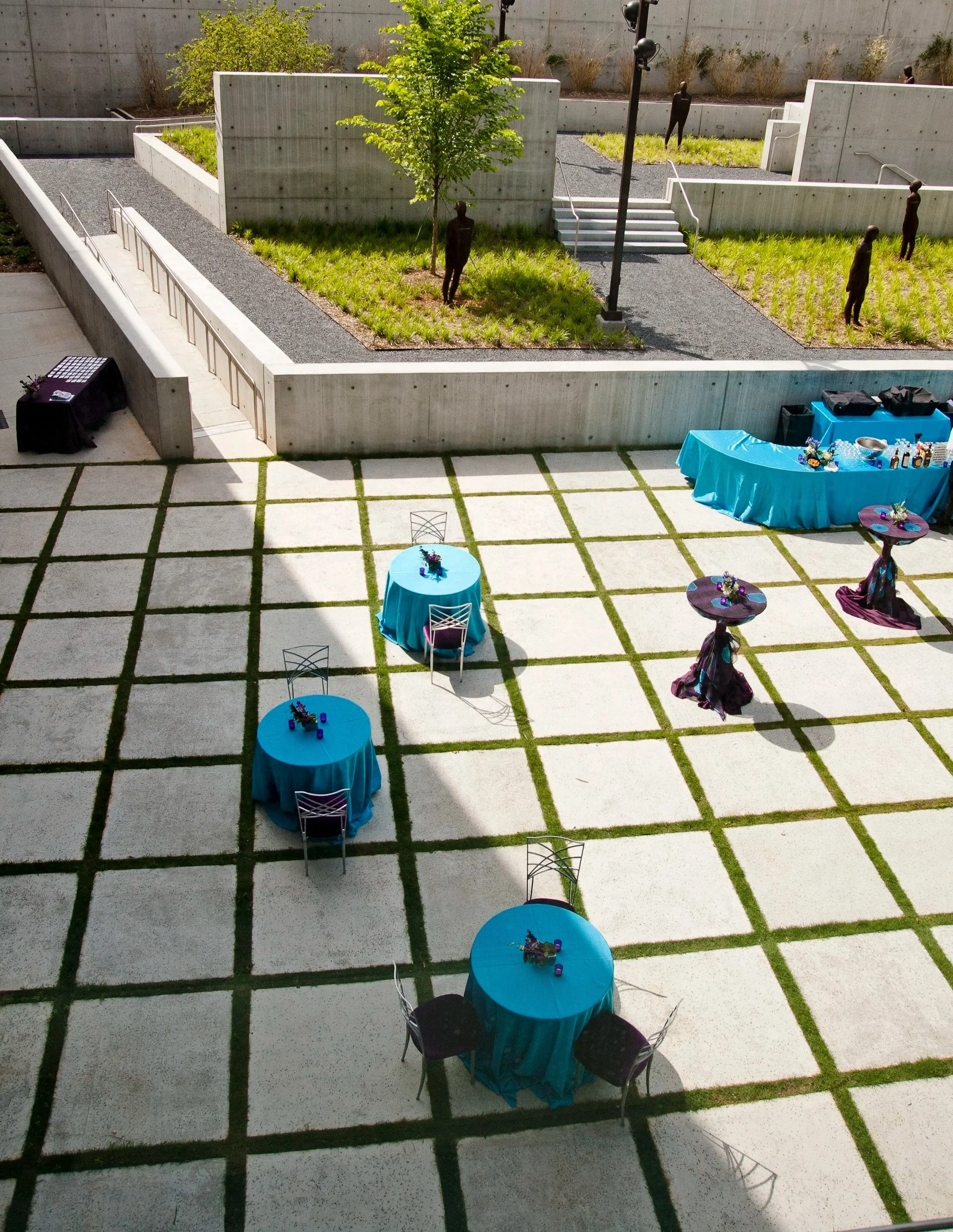A modern outdoor patio decorated for an event with round tables covered in bright blue tablecloths, small floral centerpieces, and purple decorations. There are black chairs around the tables, and a long table with a blue tablecloth holding drinks an