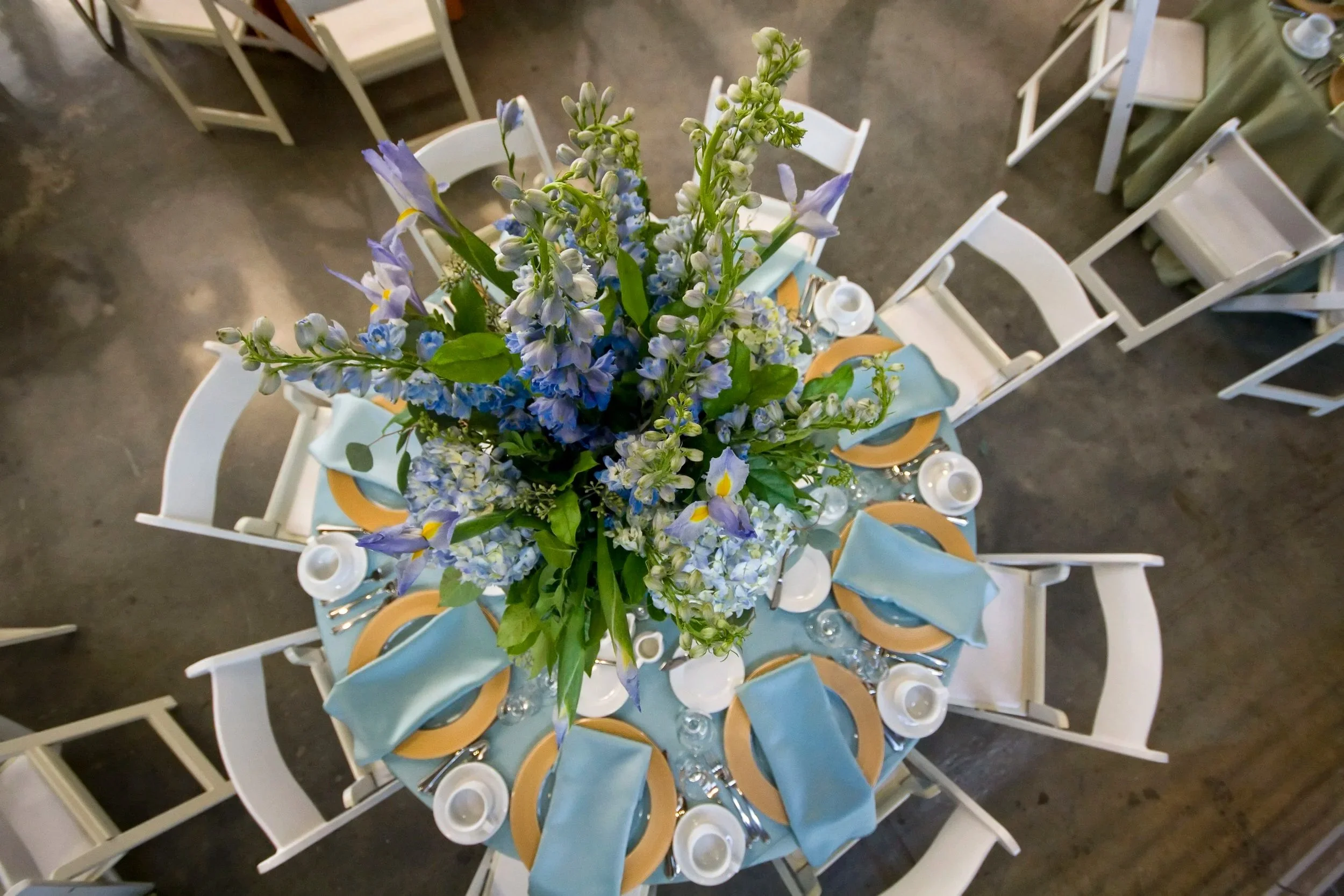 A top-down view of a round dining table decorated with a bouquet of mixed blue and white flowers, light blue cloth napkins, orange plates, and white centerpieces, surrounded by white chairs.