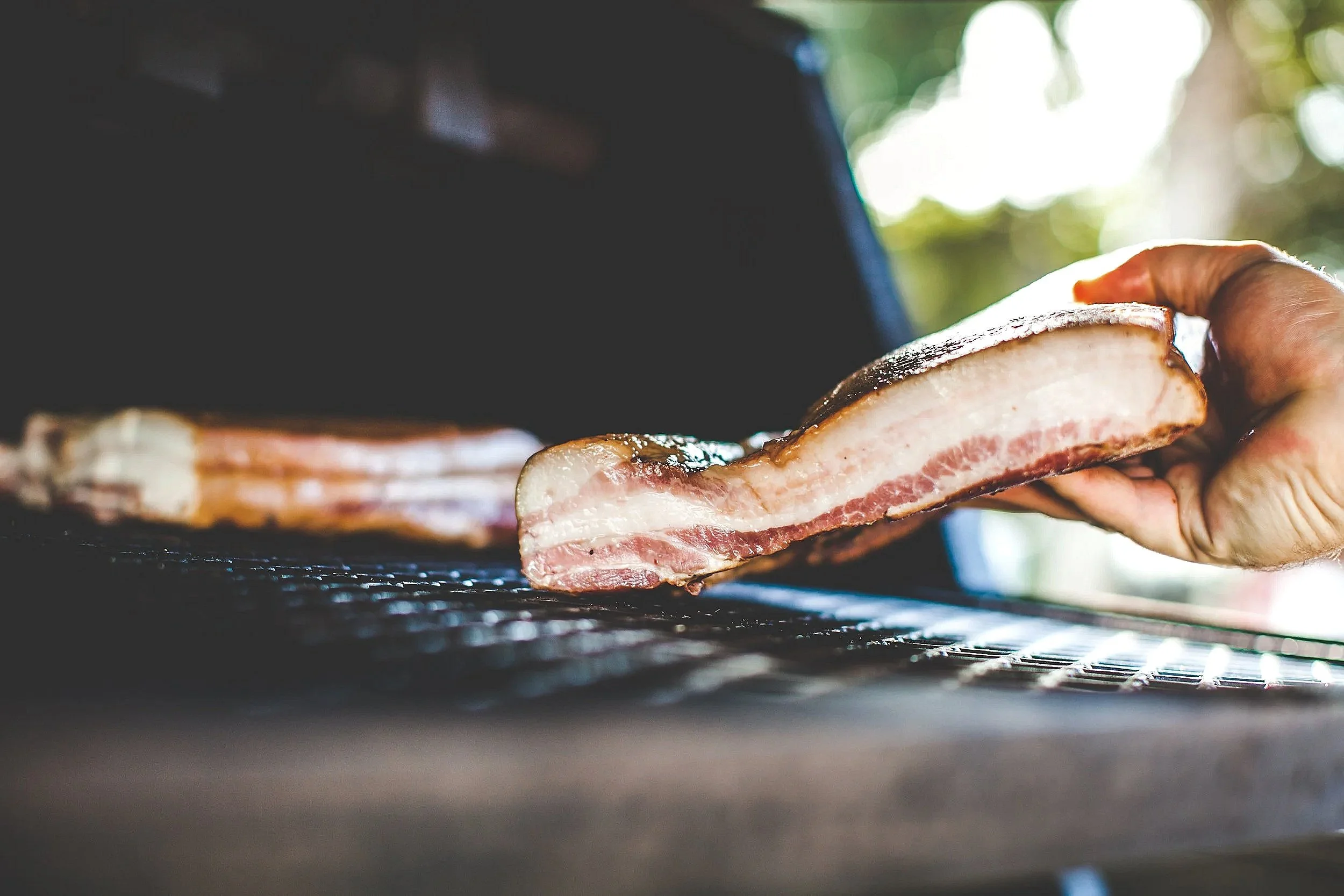 Close-up of a hand placing slices of bacon on a barbecue grill with blurred background of trees and sunlight.
