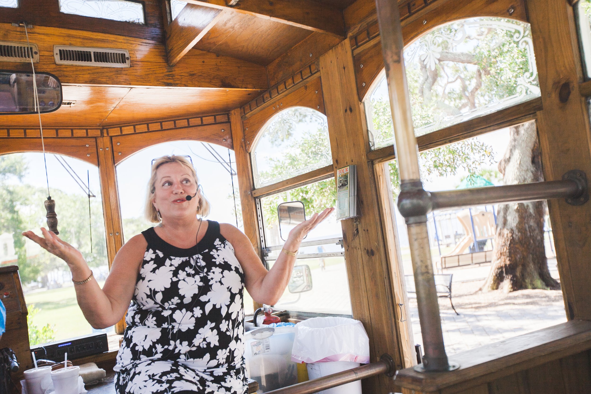 Woman with blonde hair wearing glasses, a black and white floral dress, and a headset, gesturing with her hands inside a wooden structure with large windows showing trees outside.