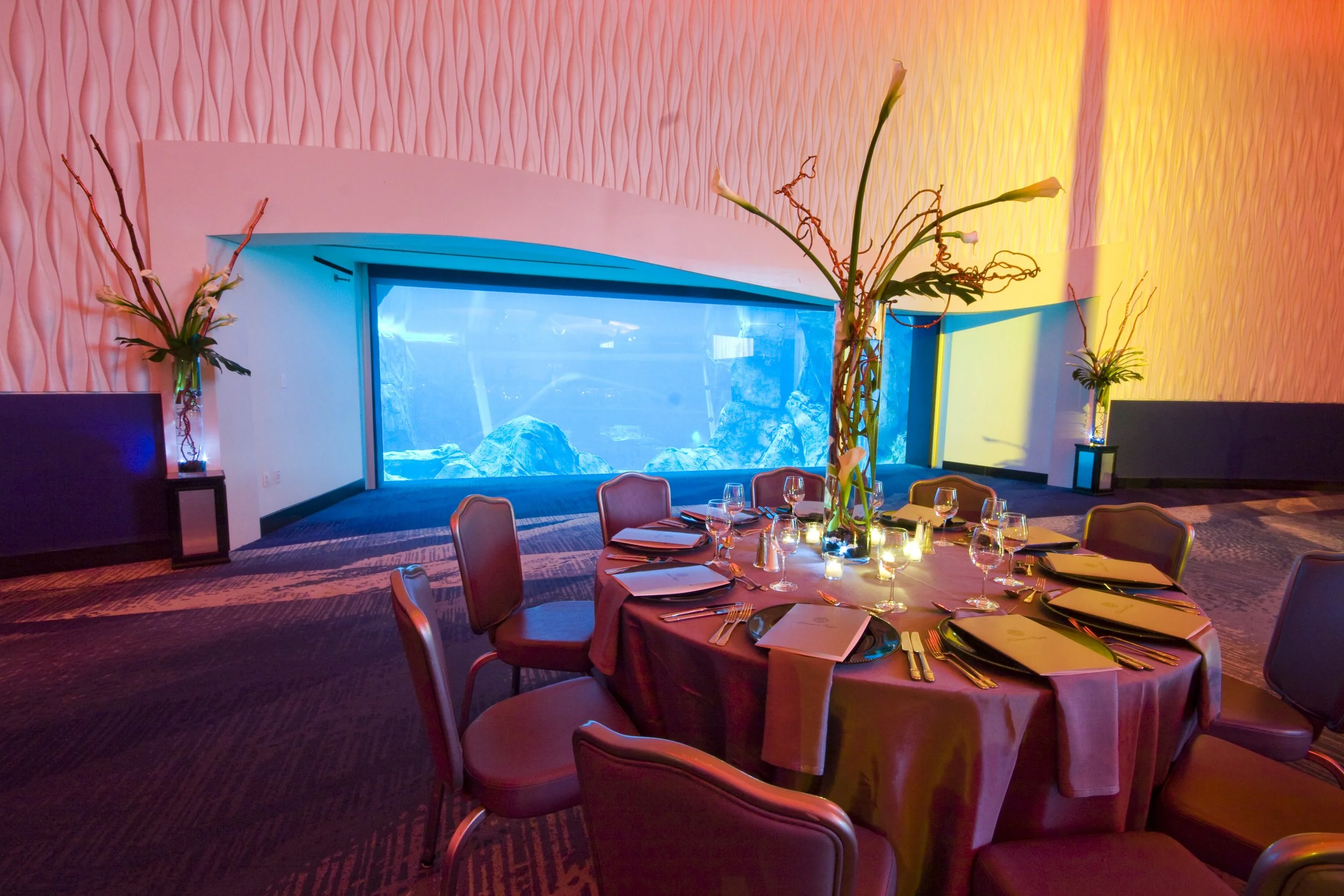 A round dining table set for a meal with wine glasses and napkins, decorated with tall flower arrangements, in a room with pink textured wall and a large aquarium with rocks.