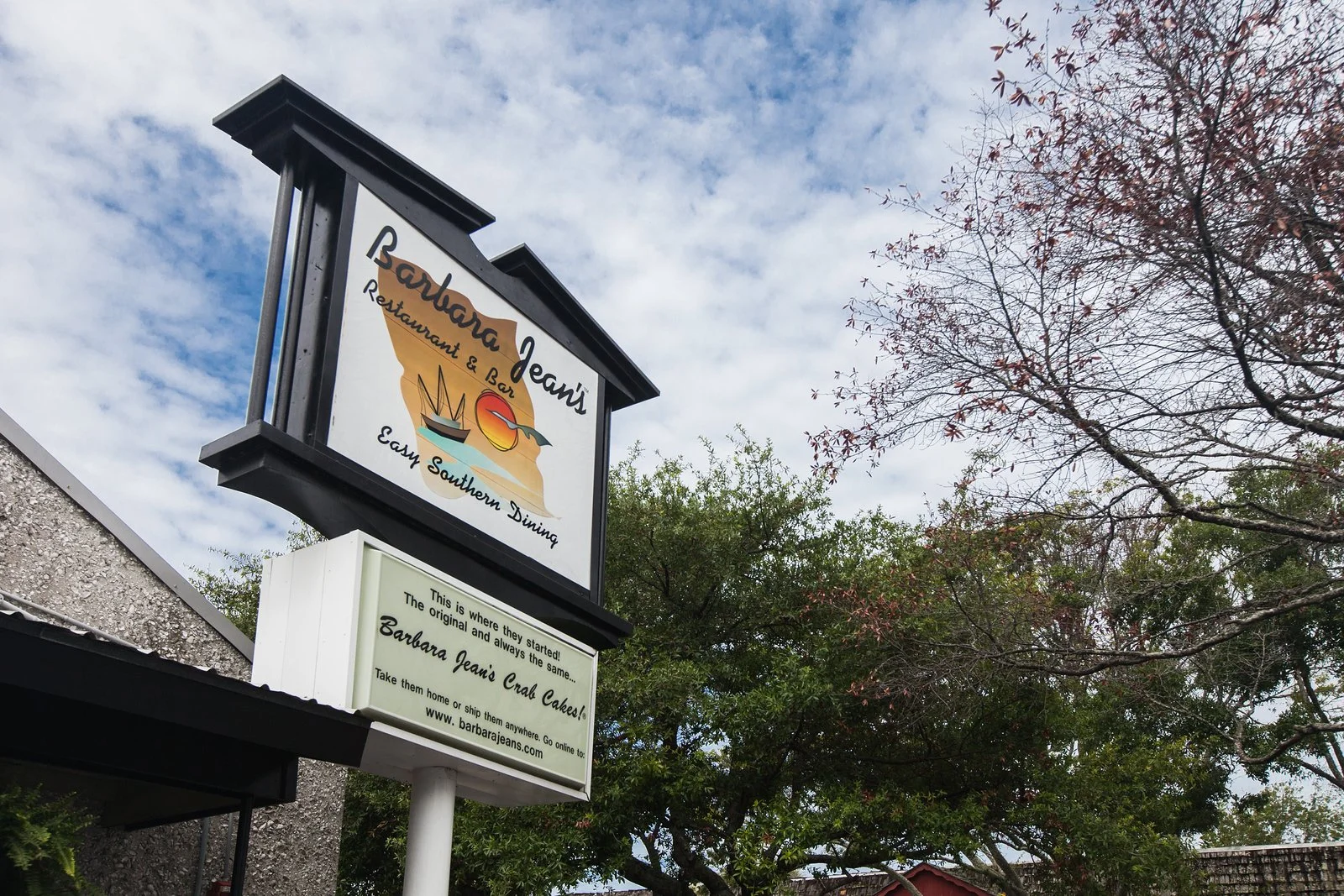 Signboard for Barbara Jean's restaurant and bar, featuring a logo with a crab, a sailboat, and a sunset, with a smaller sign below describing the restaurant's history and website.