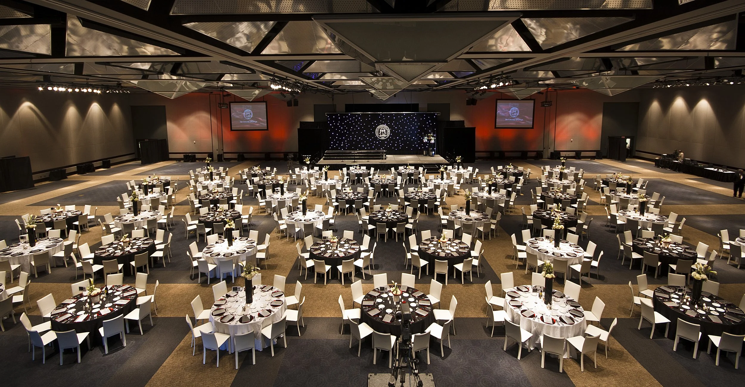 A large banquet hall set up for a formal event with round tables covered with white tablecloths, decorated with flowers, and arranged with place settings. A stage with a backdrop and large screens is at the front of the room, illuminated with soft li