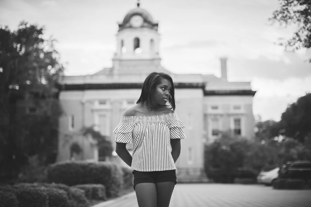 A young woman stands outside in front of a historic building with a clock tower, wearing an off-the-shoulder striped top and shorts, looking to the side.