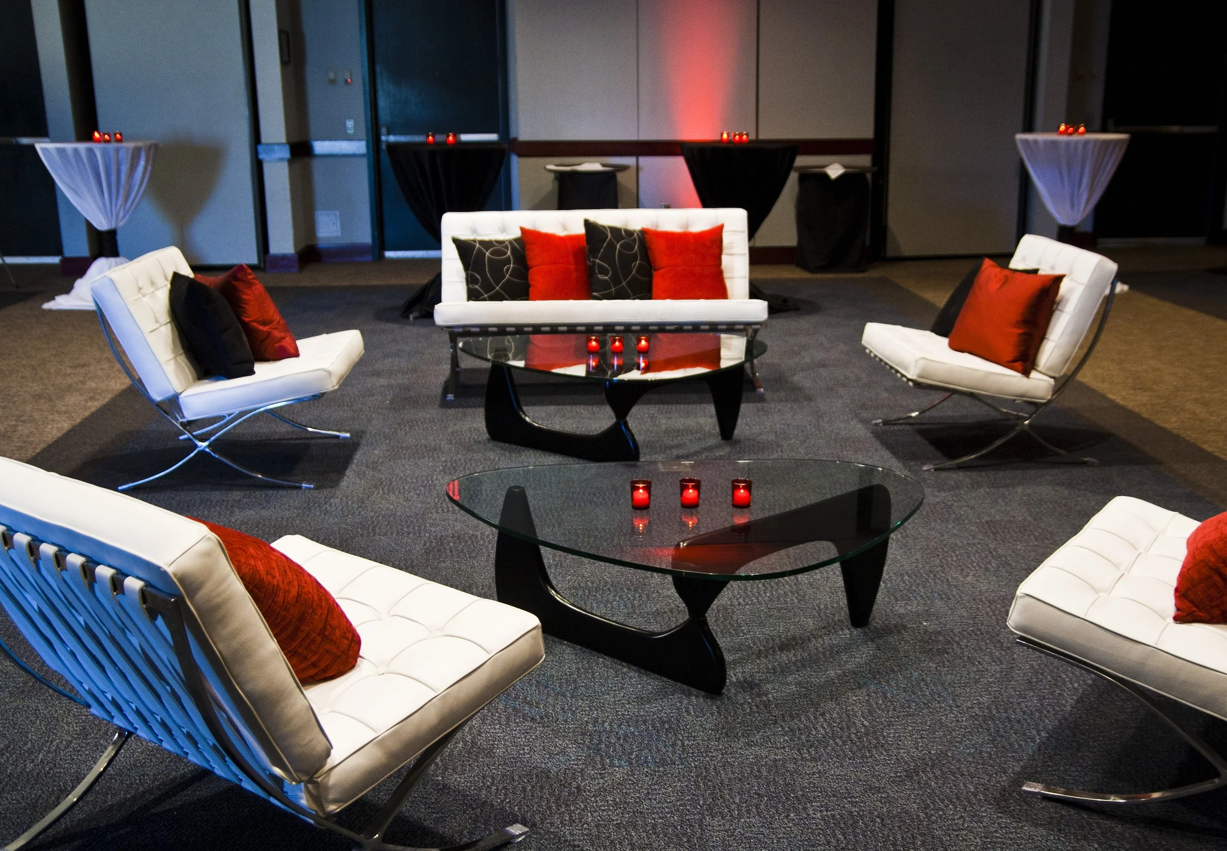 A modern lounge area with white chairs and sofas adorned with red and black cushions, glass tables with red candles, and a background of closed doors and partitions, illuminated by red lighting.
