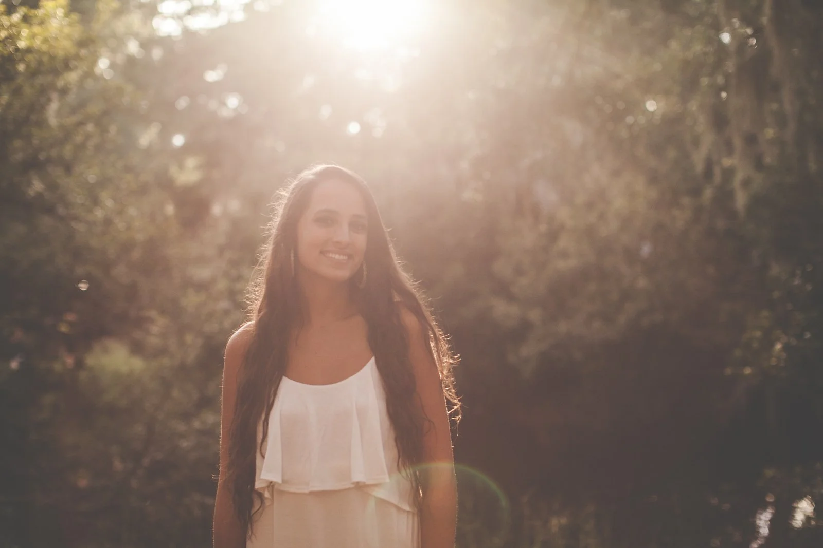 Young woman in white dress standing in golden sunlight through trees