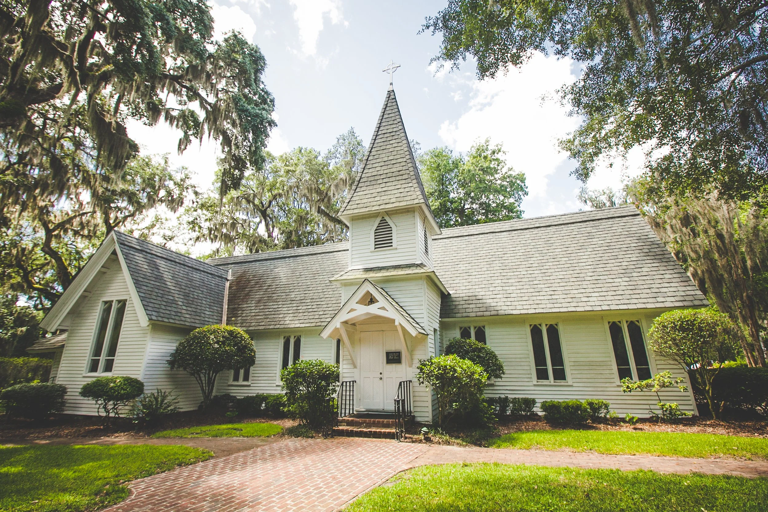 A white wooden church with a tall steeple topped with a cross, surrounded by trees with hanging moss and bushes, under a blue sky with scattered clouds.