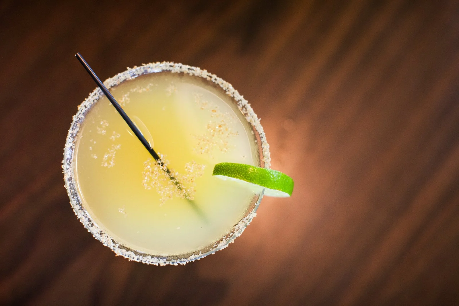 Top view of a margarita glass with salt rim, lime wedge garnish, a black straw, and a yellow cocktail inside on a brown surface.