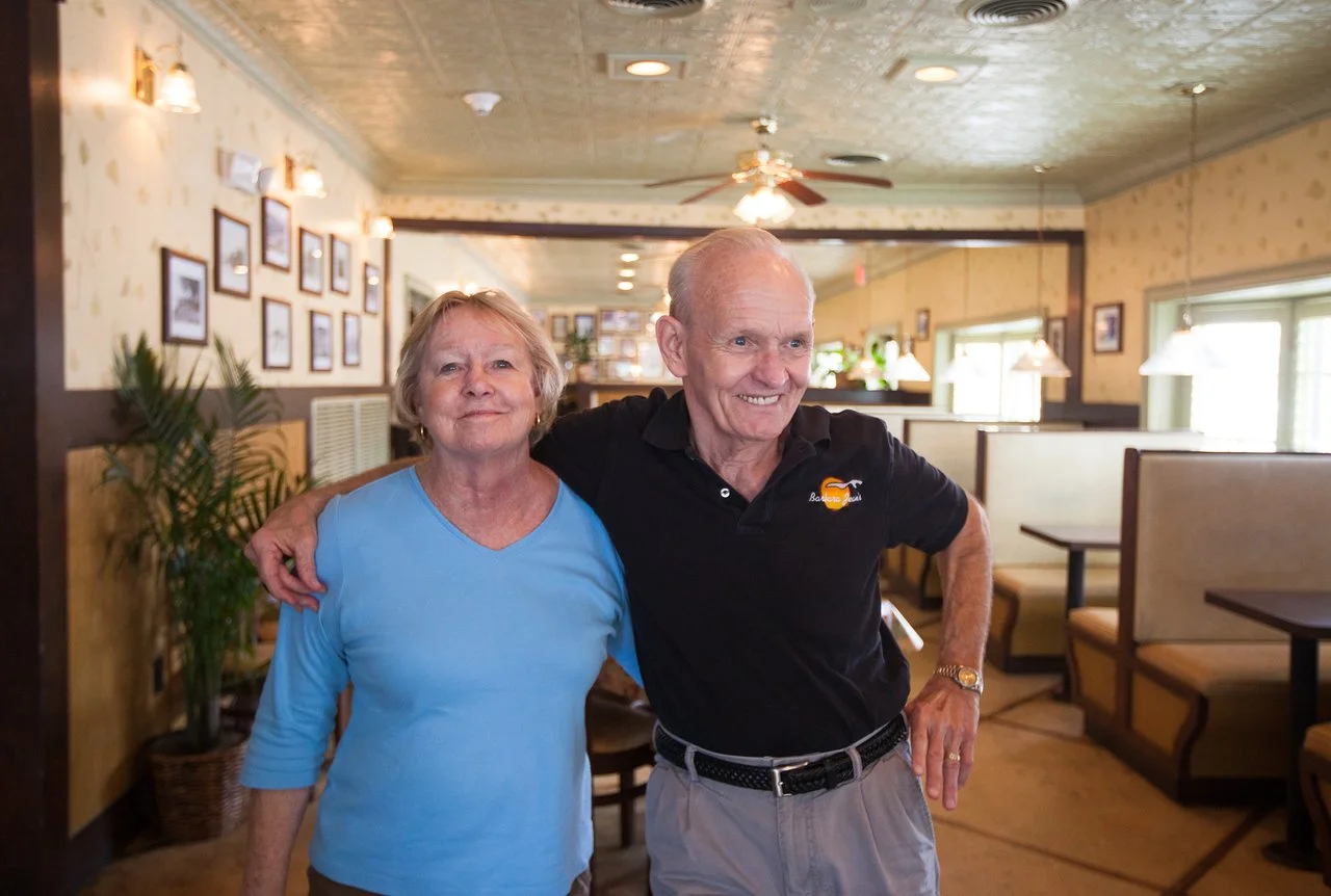 An elderly man and woman smiling and posing with their arms around each other inside a cozy restaurant. The man is wearing a black polo shirt with a logo and light-colored pants, while the woman is dressed in a light blue top. The restaurant has warm