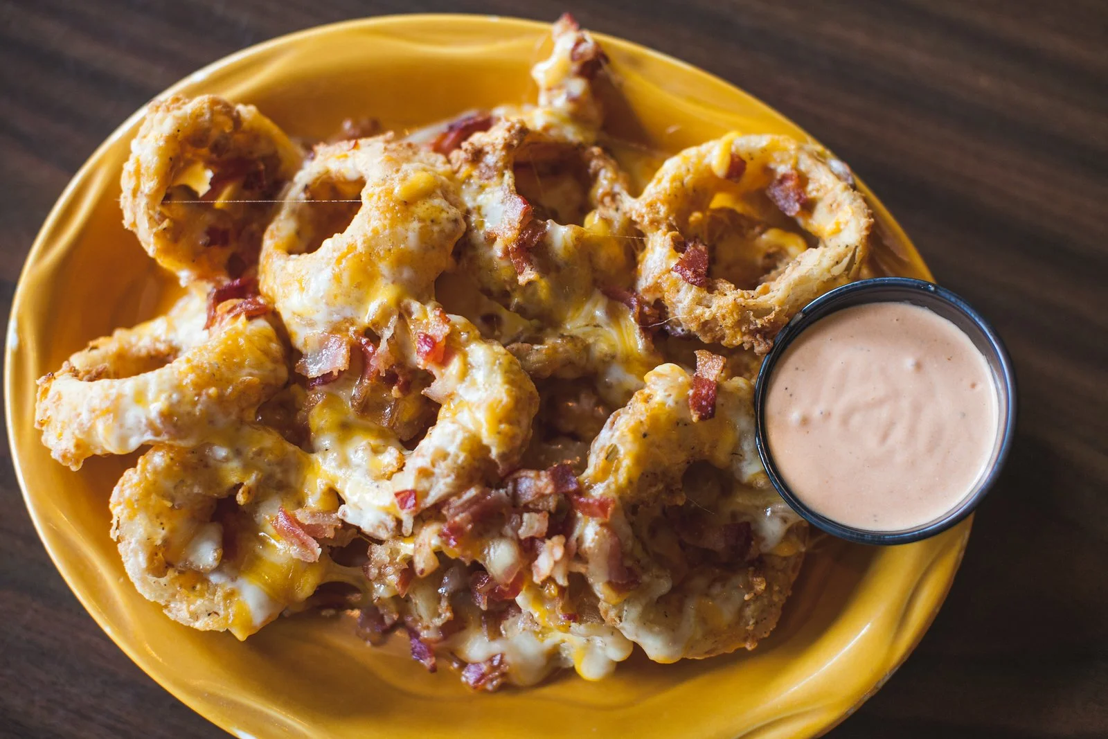 Bowl of cheese, bacon, and onion stuffed fried jalapeno poppers with a side of dipping sauce on a yellow plate