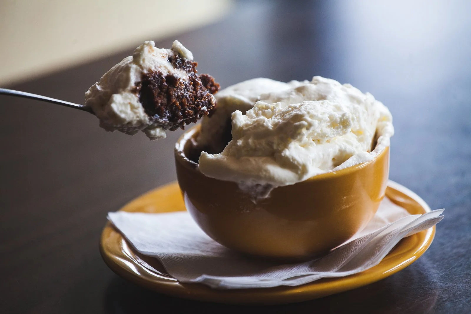 A spoonful of chocolate chip cookie dough ice cream with visible cookie chunks, held over a bowl of vanilla ice cream in a yellow bowl on a matching yellow plate with a white napkin underneath, on a dark surface.