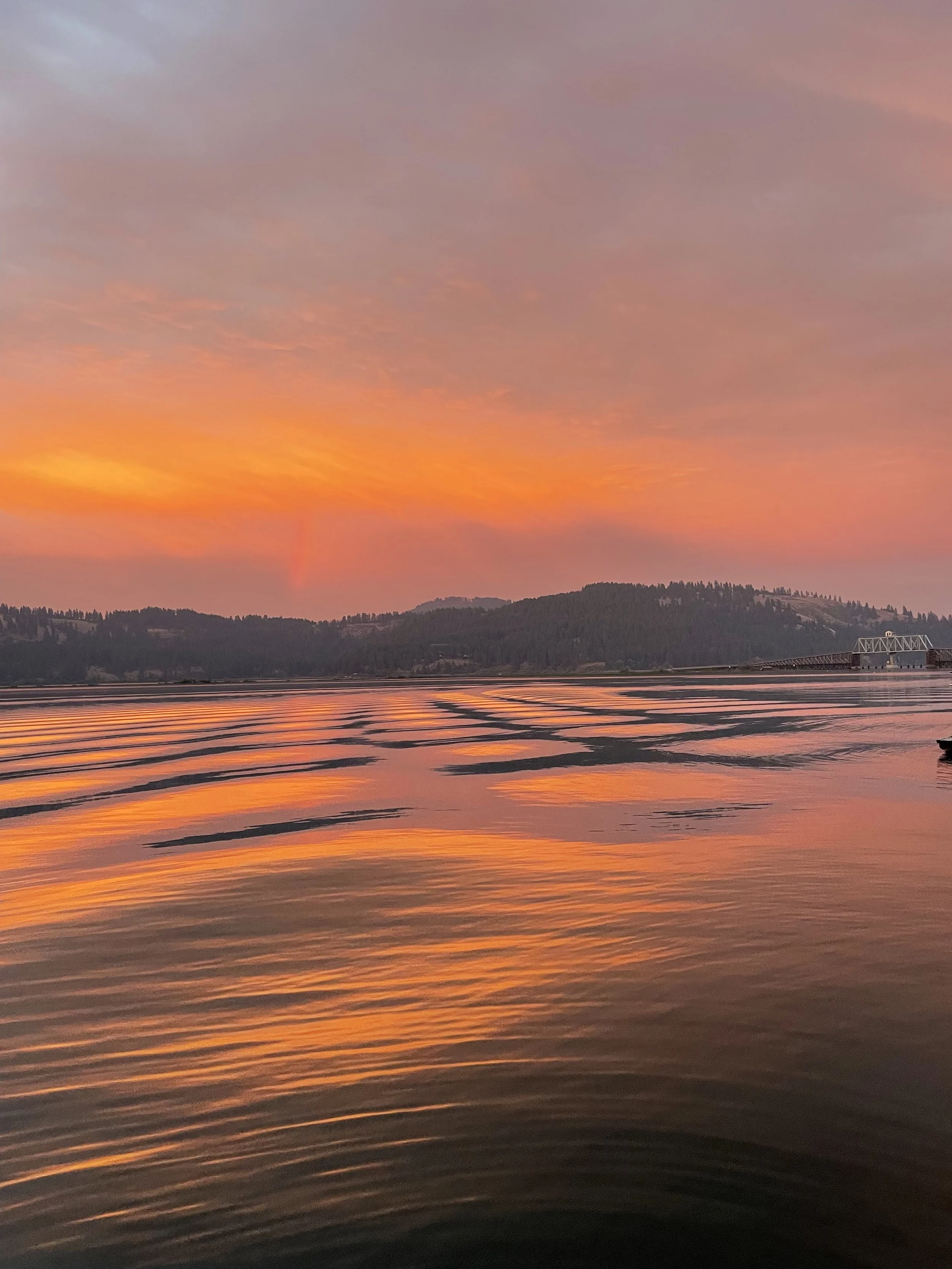 Sunset over a body of water with silhouettes of hills in the background and ripples on the water's surface.