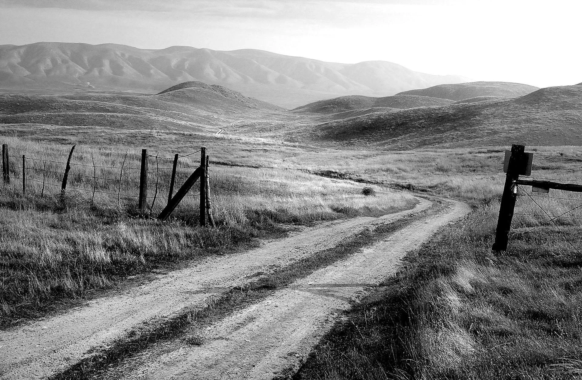 Black and white photo of a rural landscape with a dirt road winding through grassy fields, supported by a fence on the left and a broken gate on the right, with rolling hills and distant mountains in the background.