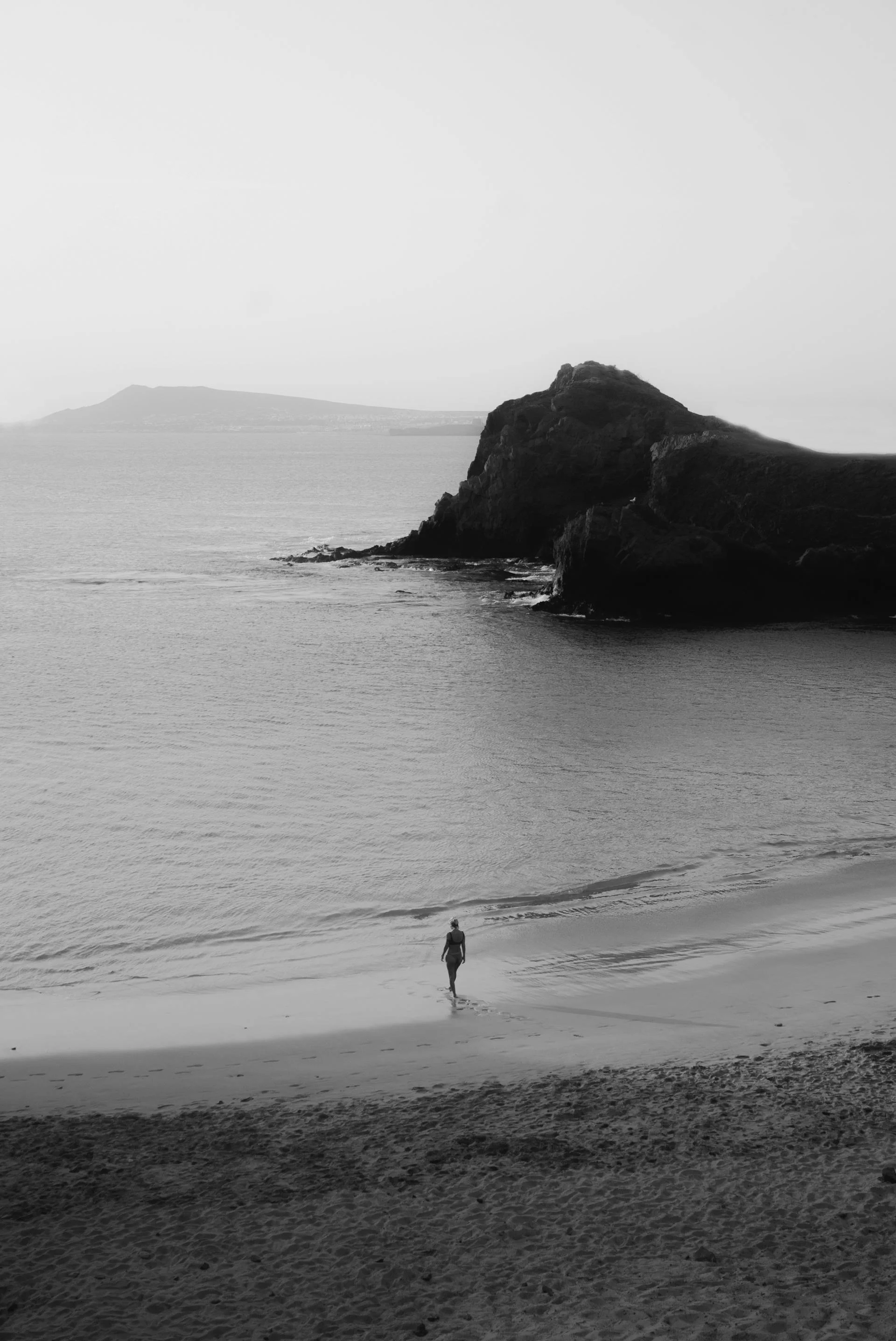 A person walking alone on a sandy beach toward the water with rocky cliffs and distant landmass in the background, black and white photo.