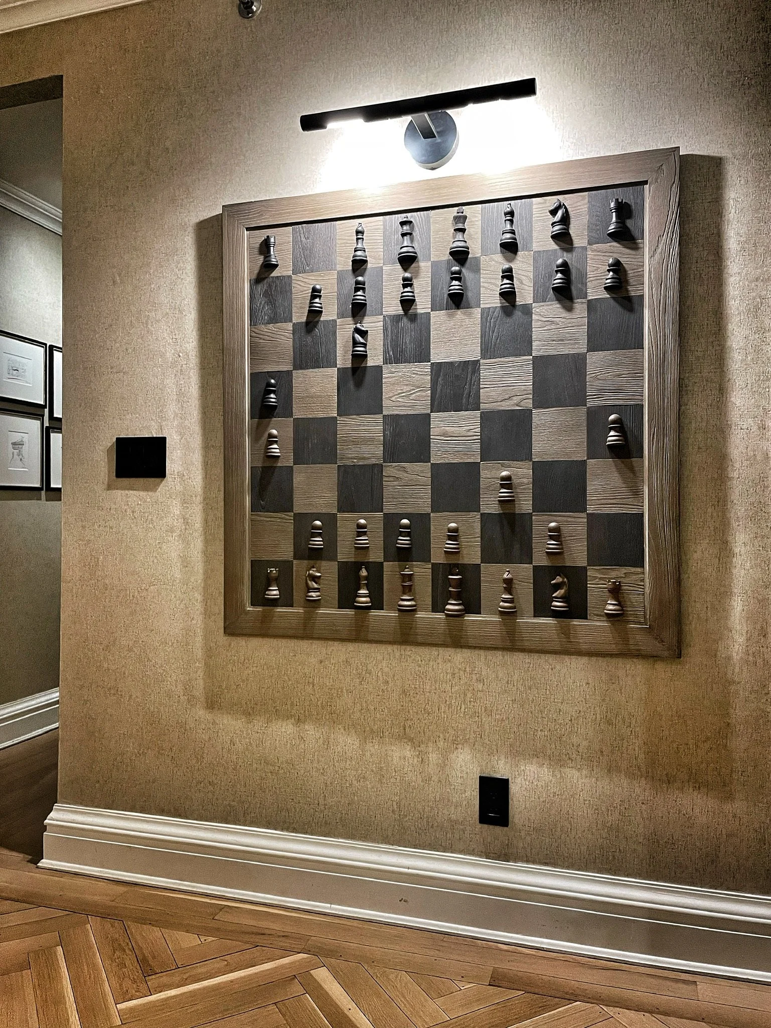 A large chessboard wall art with 3D chess pieces on a wall in a room with beige wallpaper and wooden flooring. Above the art is a black modern light fixture.