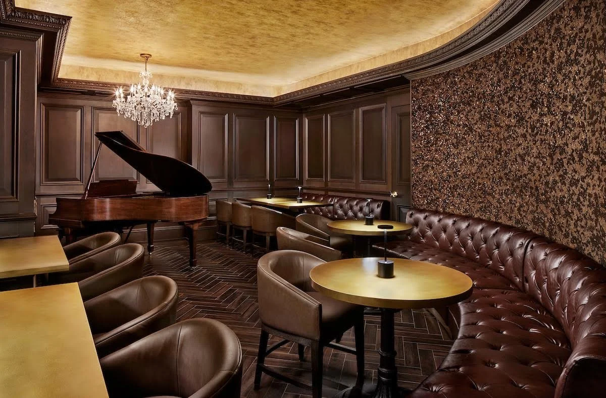 A cozy lounge with chocolate brown leather chairs and a matching banquette, gold tables, dark wood paneled walls, a grand piano, a crystal chandelier, and a patterned accent wall.