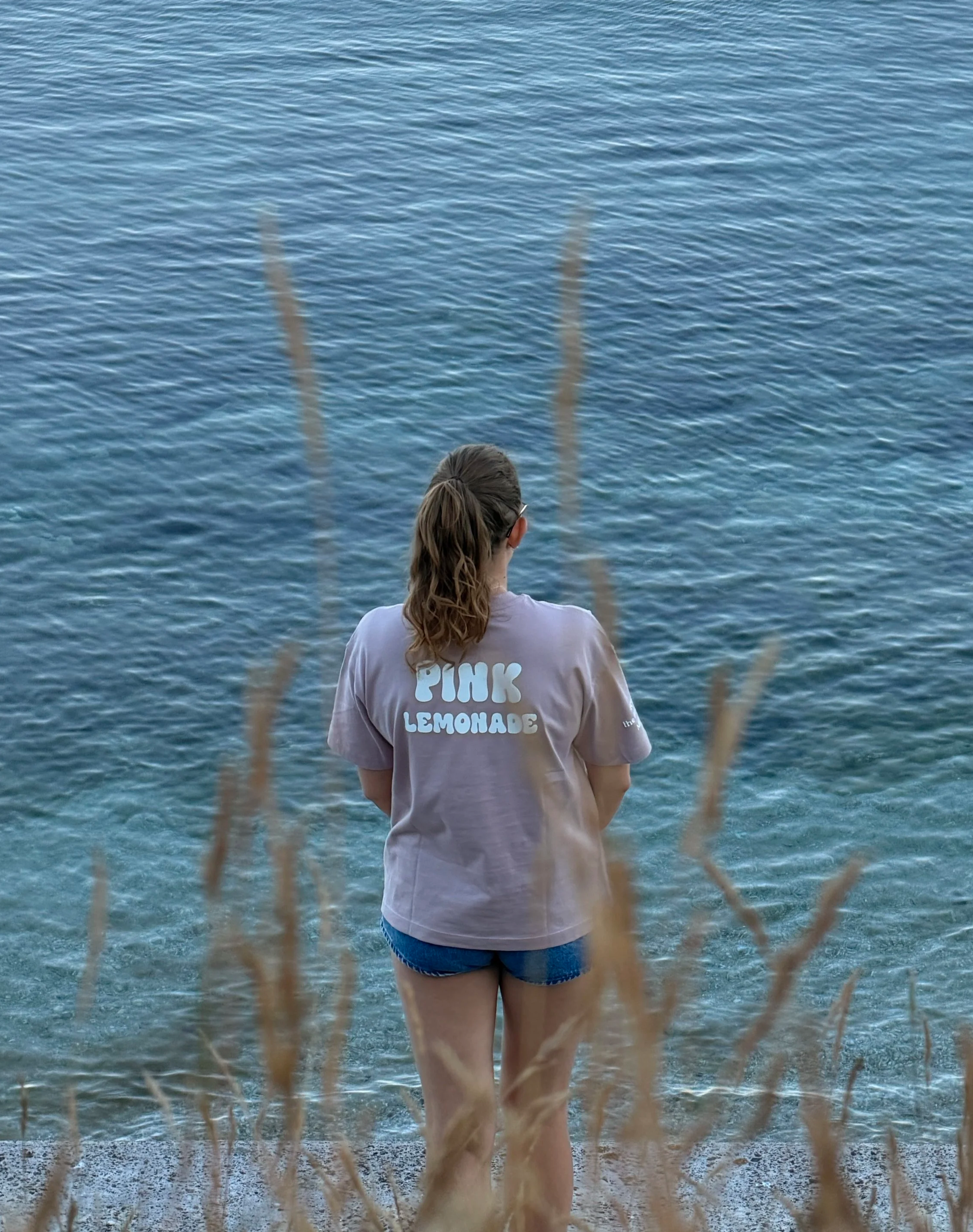 A woman with hair tied in a ponytail, wearing a light purple T-shirt with the words "PINK LEMONADE" on the back, standing near the shore of a blue body of water, viewed from behind through a fishing net.