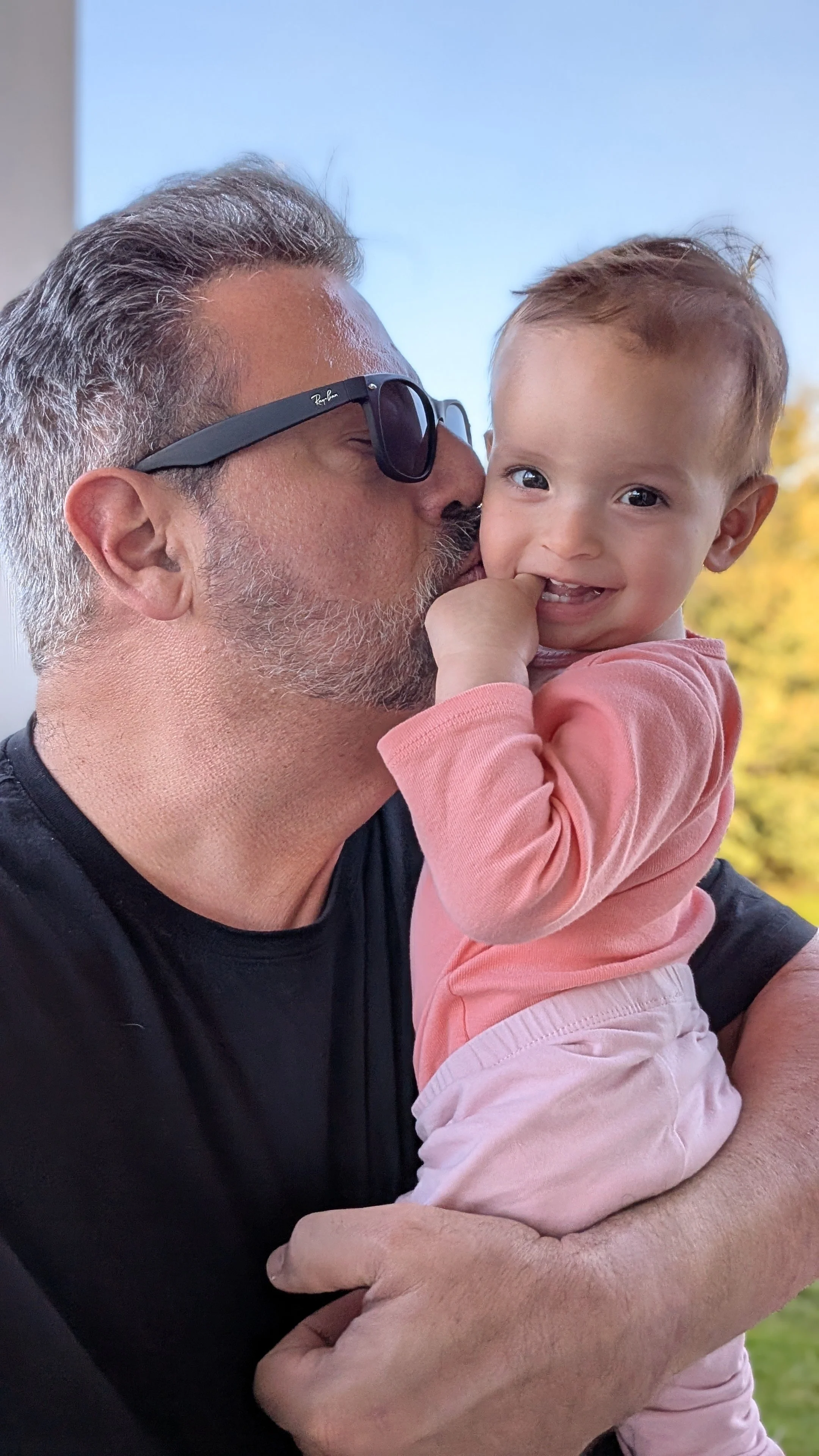 An older man with gray hair, beard, wearing sunglasses, is kissing a young girl on the cheek. The girl has light brown hair, is wearing a pink long-sleeve shirt, and is smiling while holding her finger to her mouth. They are outdoors with a blue sky and some trees in the background.