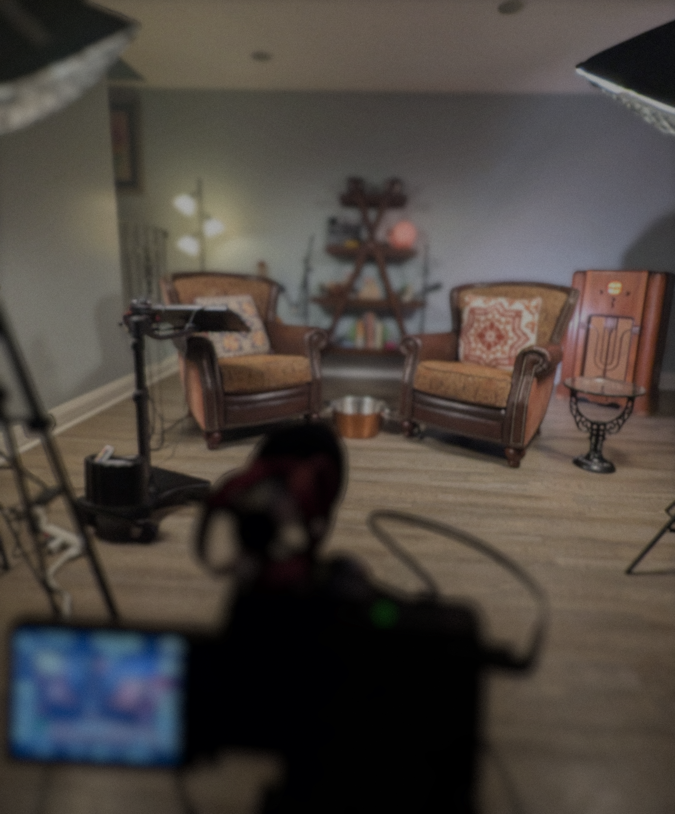 A room with wooden furniture including two chairs with patterned cushions, a table, and a bookshelf with decorations. There are wall lights, and a small framed picture on the wall. The photo appears blurry.