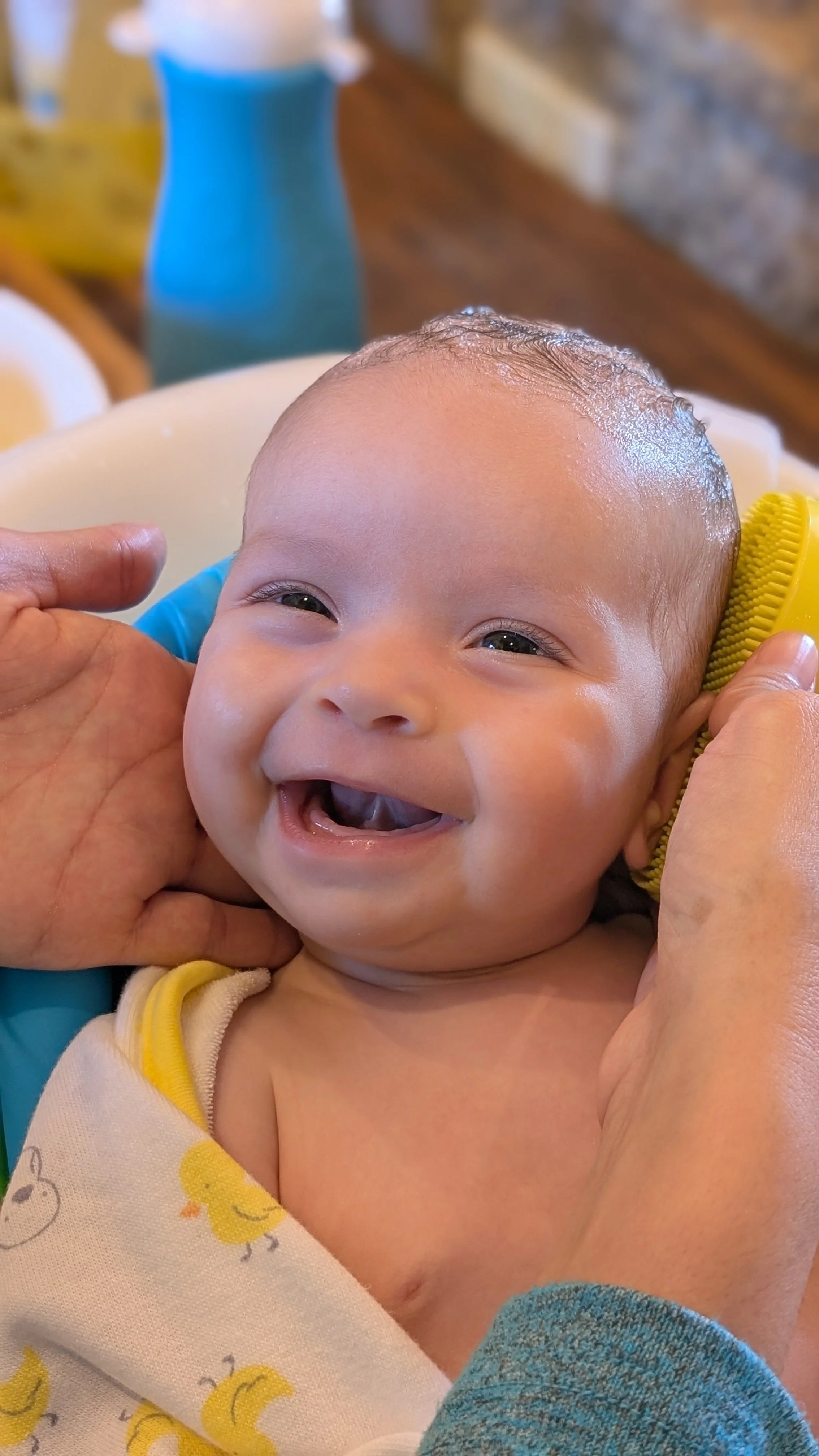 Close-up of a smiling baby with wet hair, lying in a bath, surrounded by adult hands gently holding the baby's face and head.