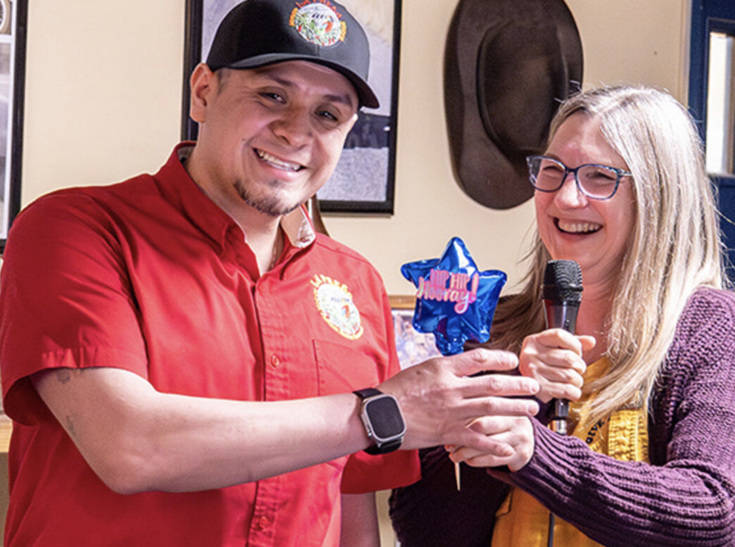A man in a red shirt and a woman with glasses and long blonde hair smiling and exchanging a small blue balloon with pink writing while holding a microphone.