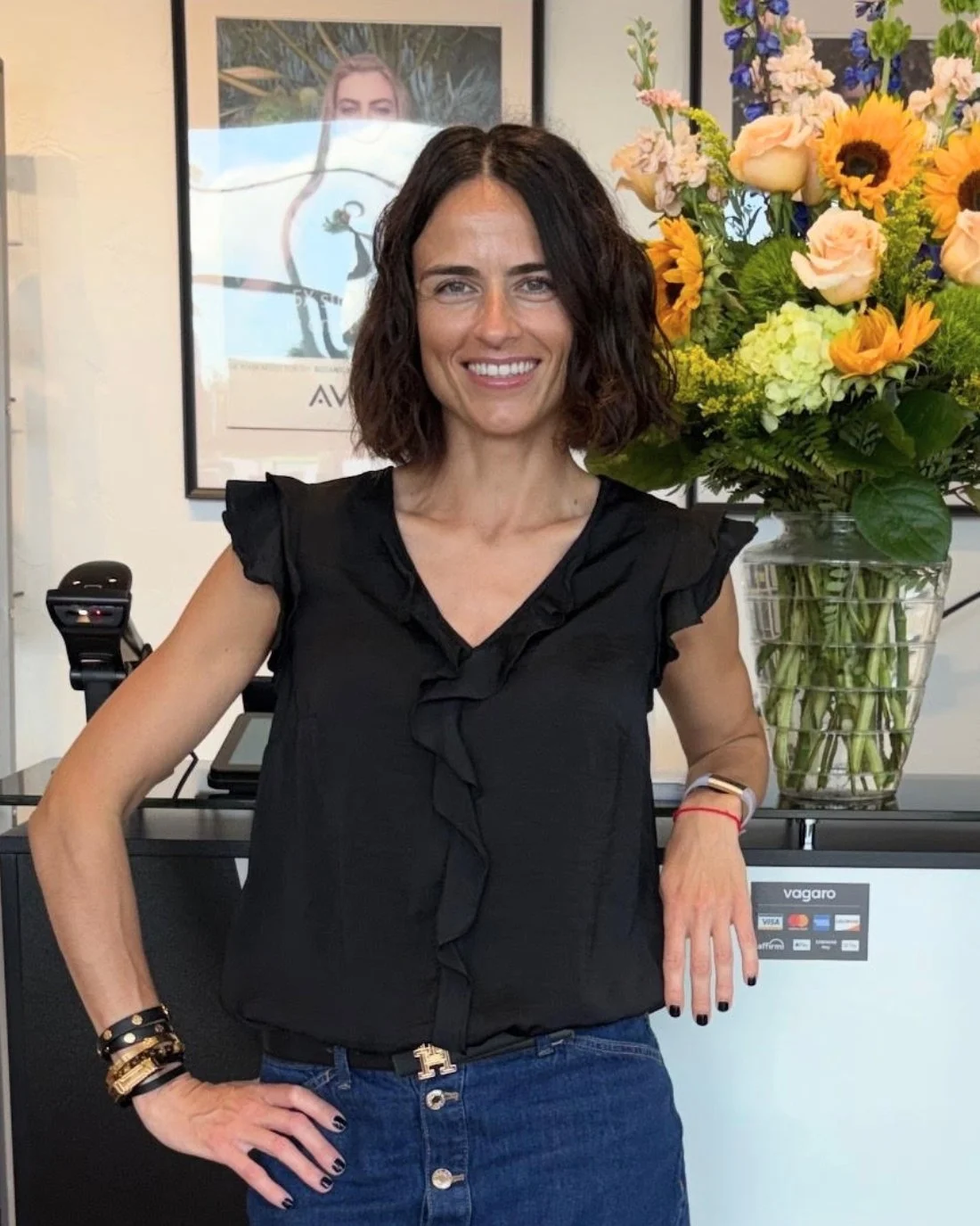 A woman with shoulder-length curly brown hair smiling, wearing a black top and jeans, standing beside a large vase of colorful flowers, in what appears to be an indoor setting.