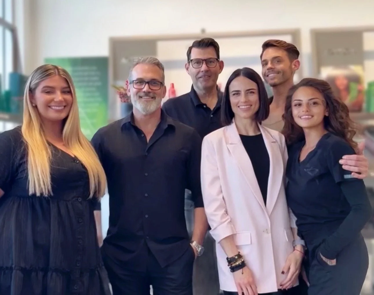 Group of six diverse professionals smiling indoors, standing close together, dressed in business casual attire.