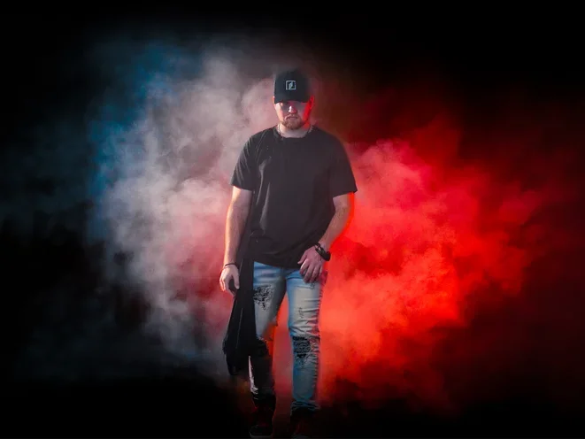 A man wearing a black cap and black t-shirt walks through colorful smoke with blue and red hues, against a dark background. Micah Ariss