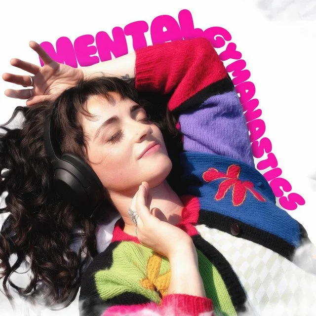 Young woman with curly dark hair and headphones lying down with eyes closed, wearing colorful cardigan with flowers, against a pink background with text 'MENTAL GYMNASTICS'. Lindsey Ponder