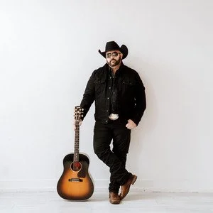 A man dressed in black, wearing a cowboy hat, sunglasses, and boots, standing next to an acoustic guitar against a plain white background. Kurt Van Meter