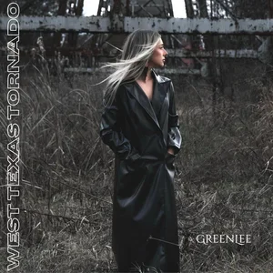 A woman with long blonde hair standing outdoors in front of a weathered fence and broken highway overpass, wearing a black leather coat. Morgan Greenlee