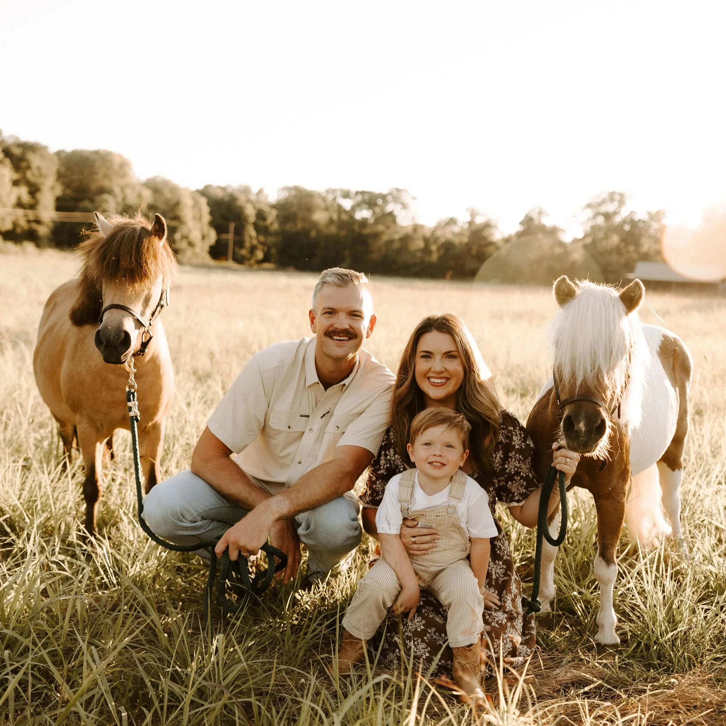 A man and woman smiling outdoors with children and horses in a field during sunset.
