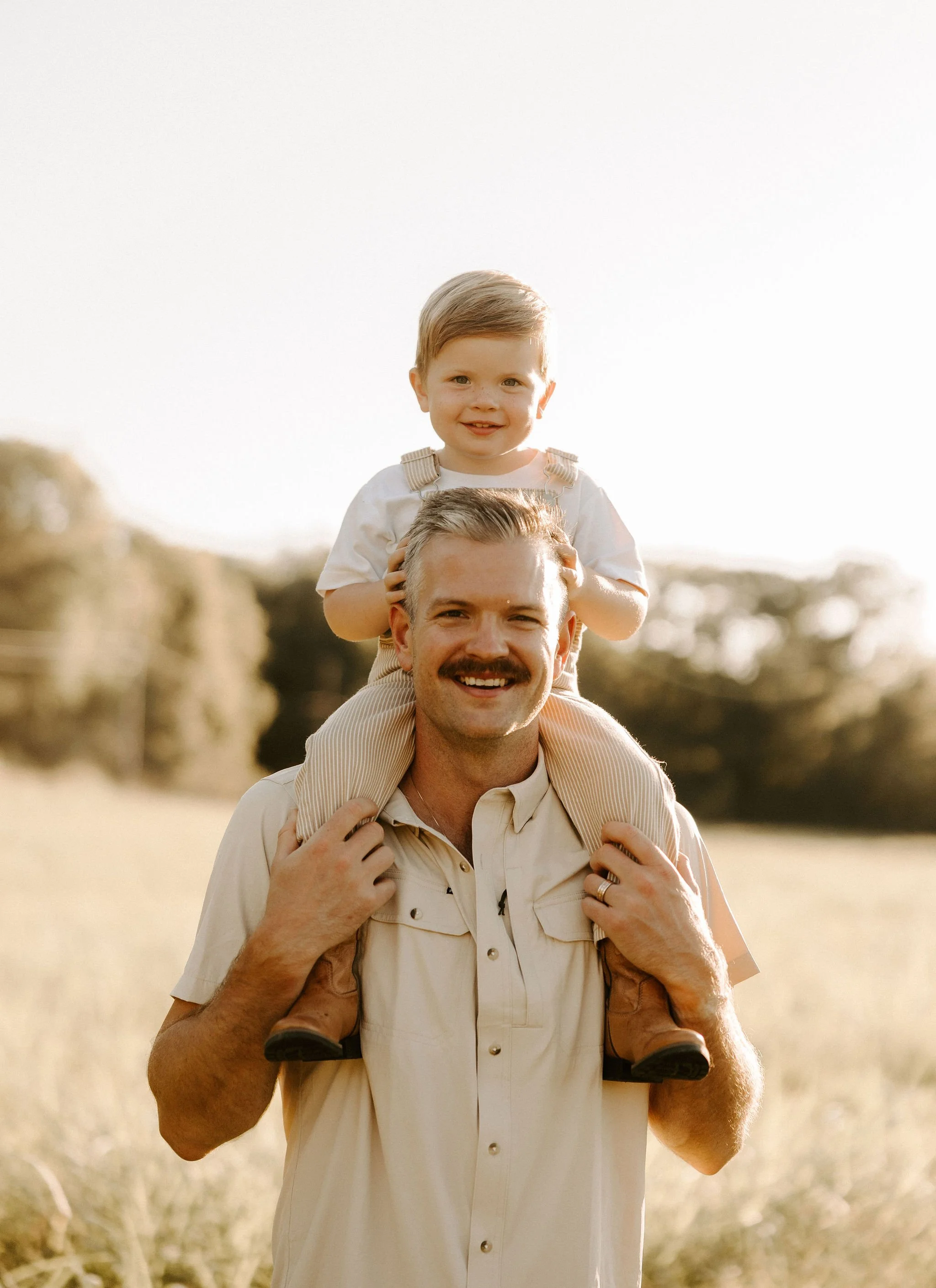 A man with a mustache carrying a young boy on his shoulders in a field during golden hour, with trees in the background.