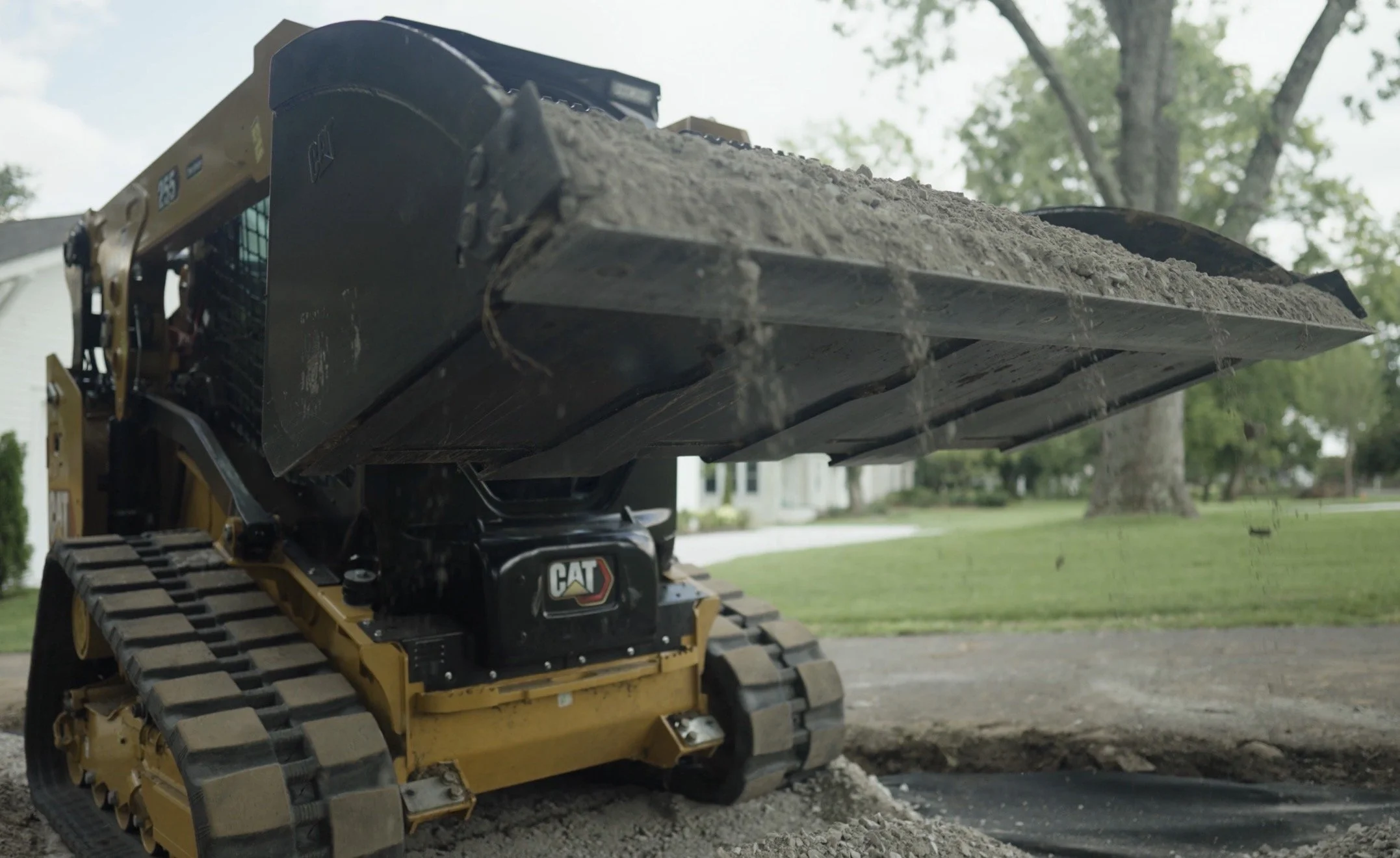 A small Caterpillar excavator with tracks is moving dirt in a residential neighborhood with trees and houses in the background.