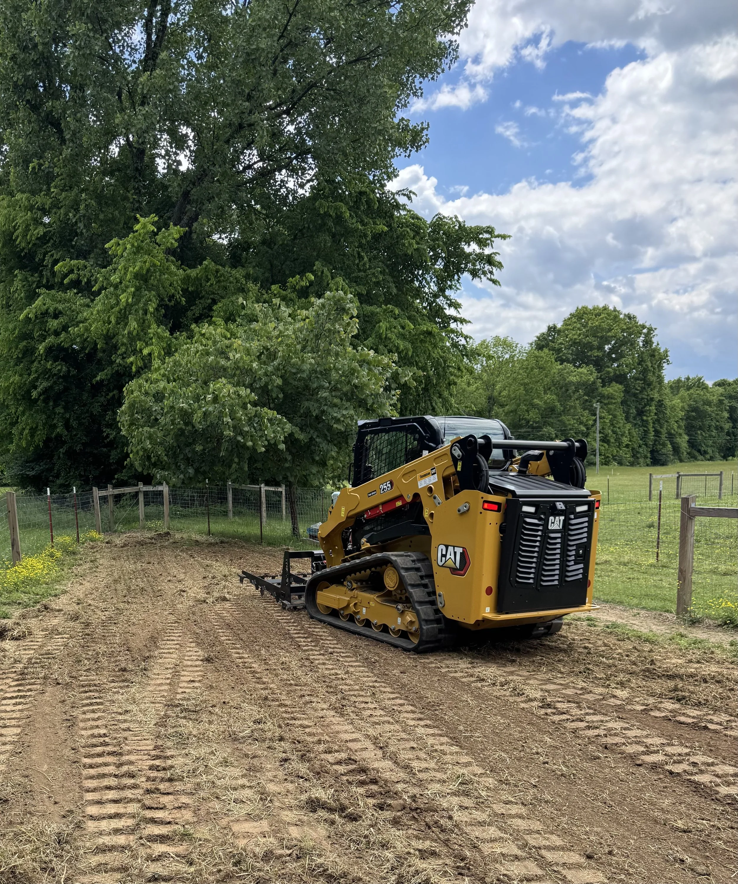 A yellow and black Caterpillar compact track loader parked on a dirt patch in a green, wooded area under a partly cloudy sky.