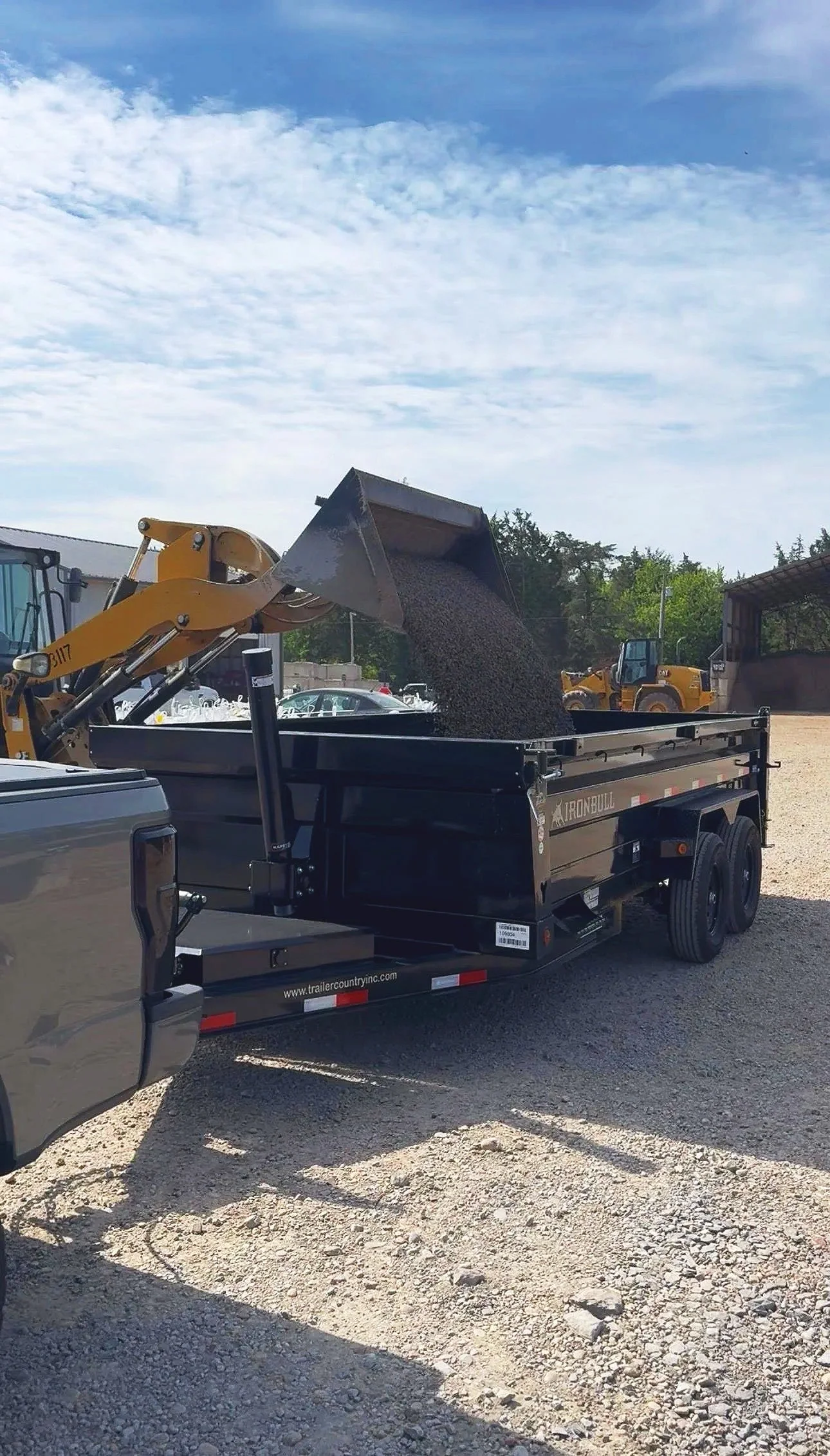 Construction site with a dump truck unloading gravel into a black trailer, yellow backhoe loader in the background, gravel ground, partly cloudy sky.