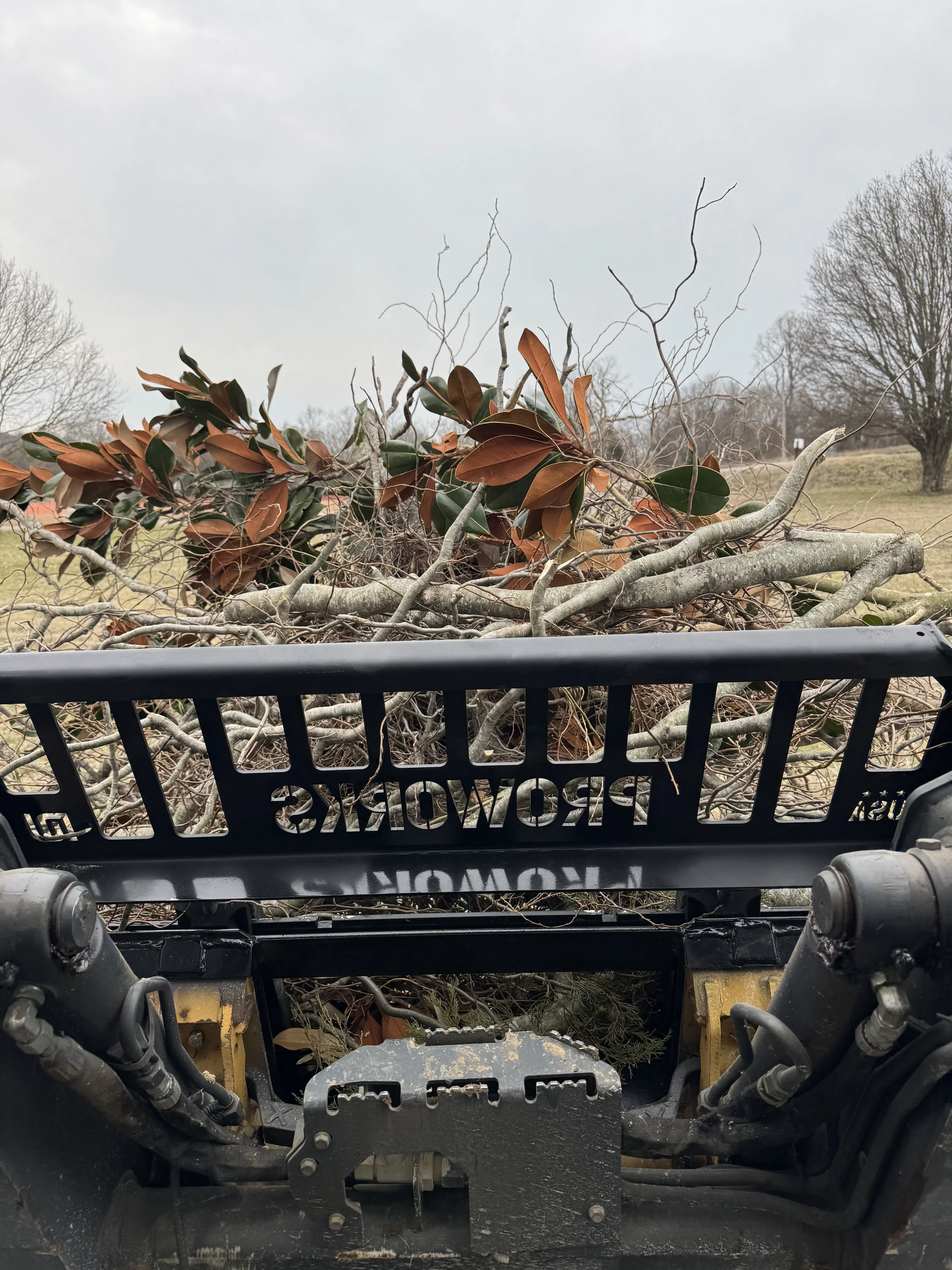 Upside-down view of a tractor's front area with parts of the engine and hydraulic system visible, behind the tractor is a pile of branches and leaves, set in an outdoor field with trees and cloudy sky.