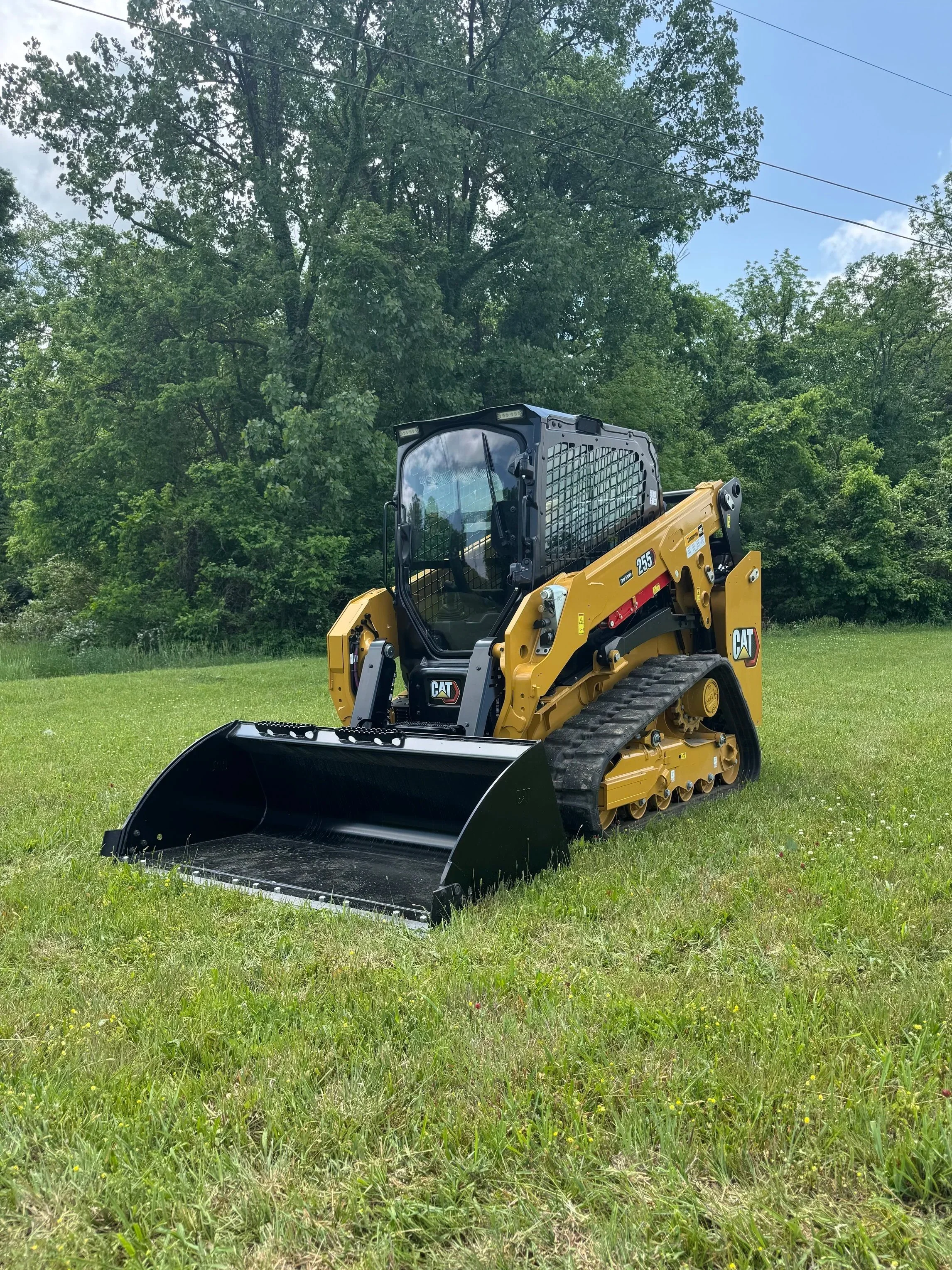 A small Caterpillar skid steer loader on a grassy field with trees and power lines in the background.