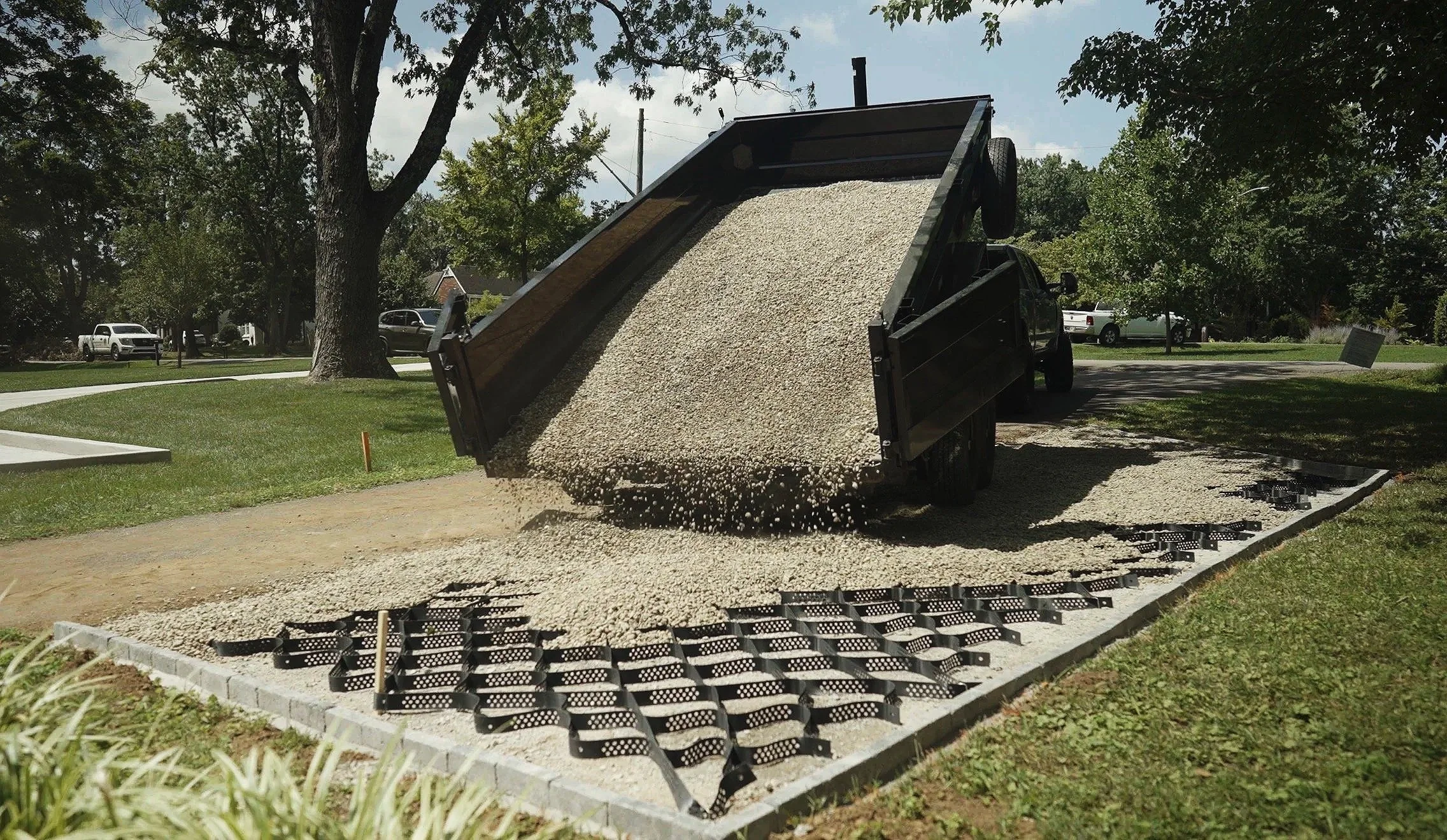 A dump truck is pouring gravel onto a foundation base during construction in a residential area with trees and parked cars visible in the background.